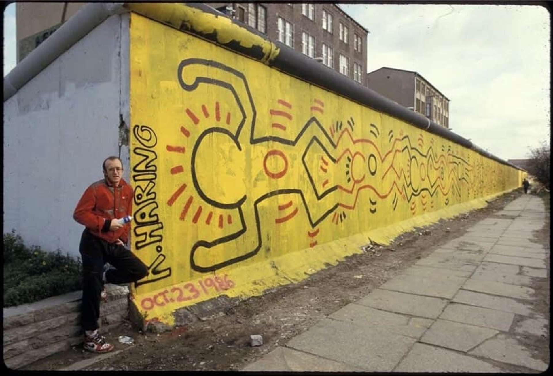 A photograph of artist Keith Haring standing in front of his mural on the Berlin Wall. The mural shows several connected figures in red and black, against a yellow background.