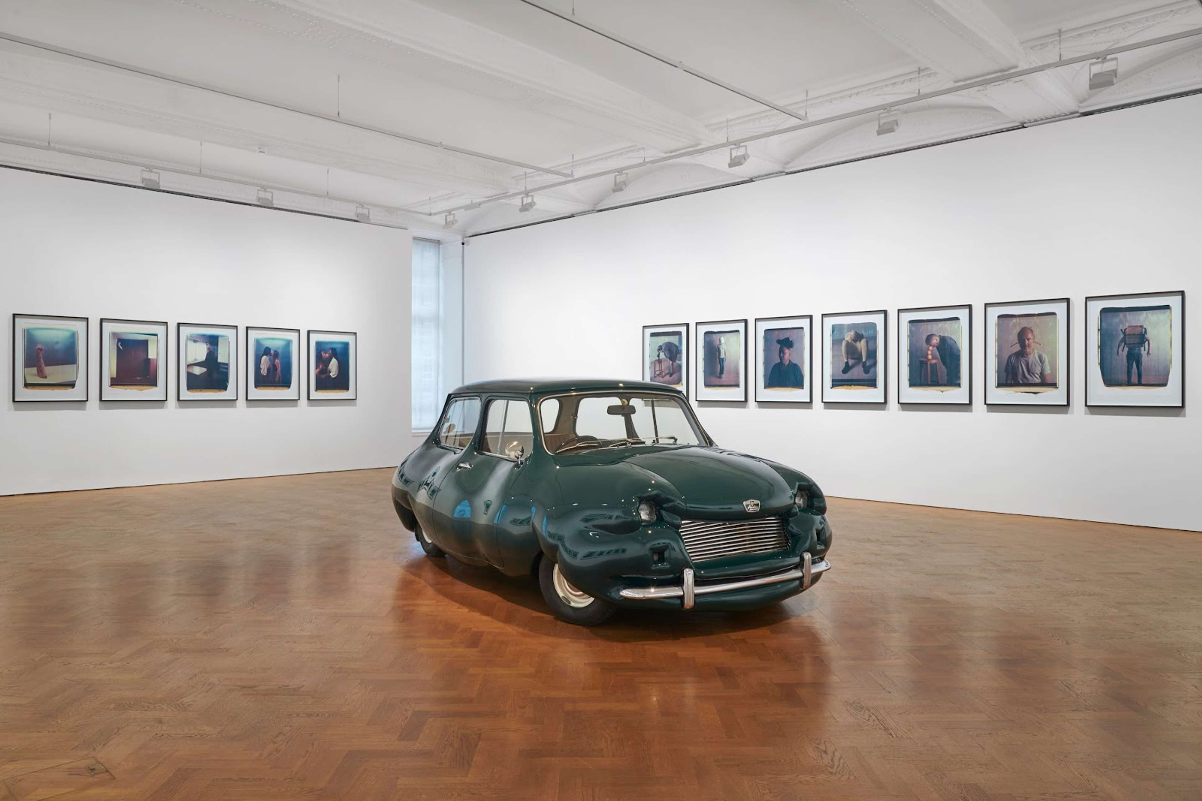 A red, fibreglass sculpture titled "Fat Mini" by Austrian artist Erwin Wurm. The sculpture is a playful representation of a classic Mini Cooper car, exaggerated in size and shape to create a plump appearance. It stands on four small wheels.