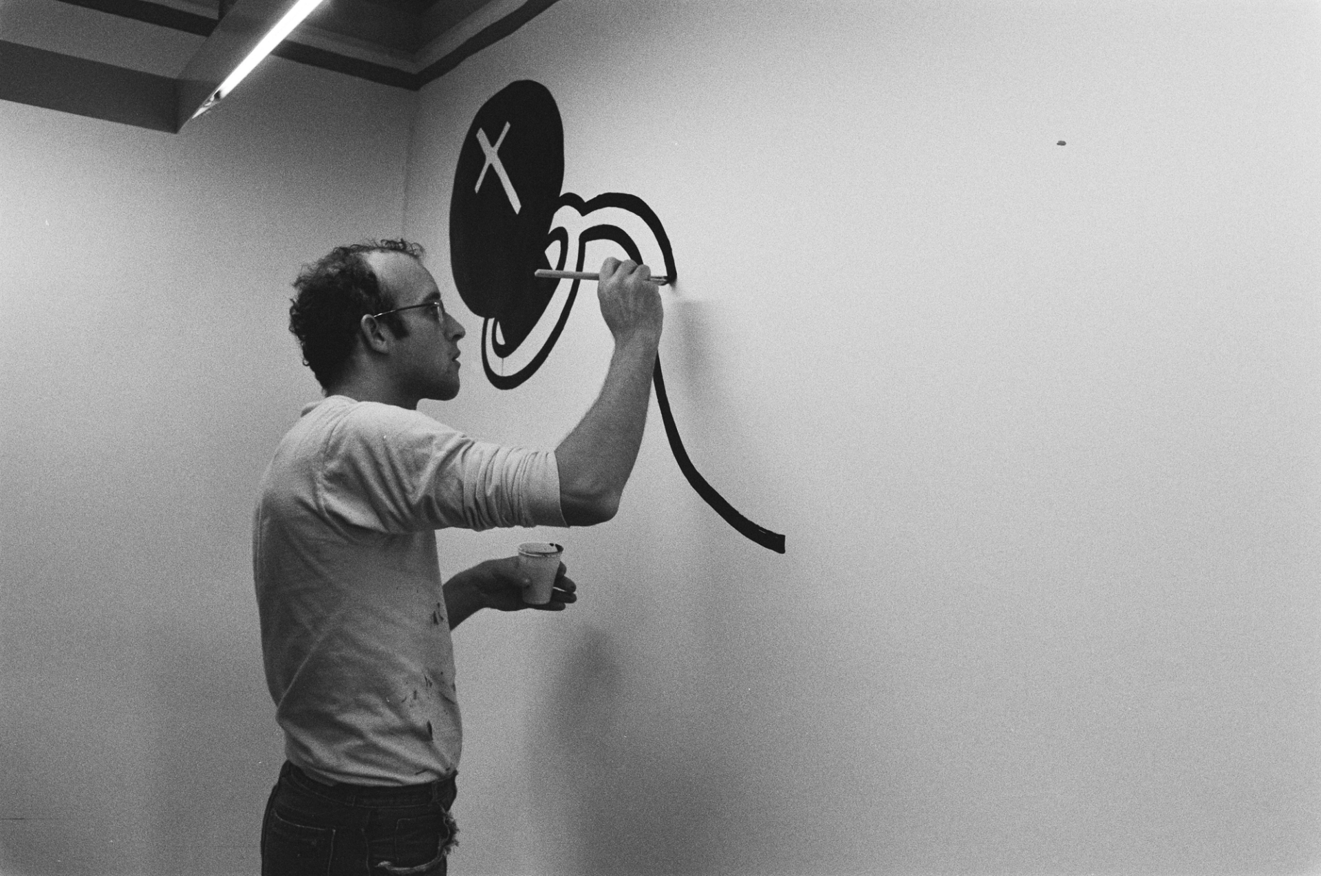 A monochrome image of the artist Keith Haring at work on a mural at the Stedelijk Museum in Amsterdam. He is wearing a long-sleeved t-shirt and his signature glasses.