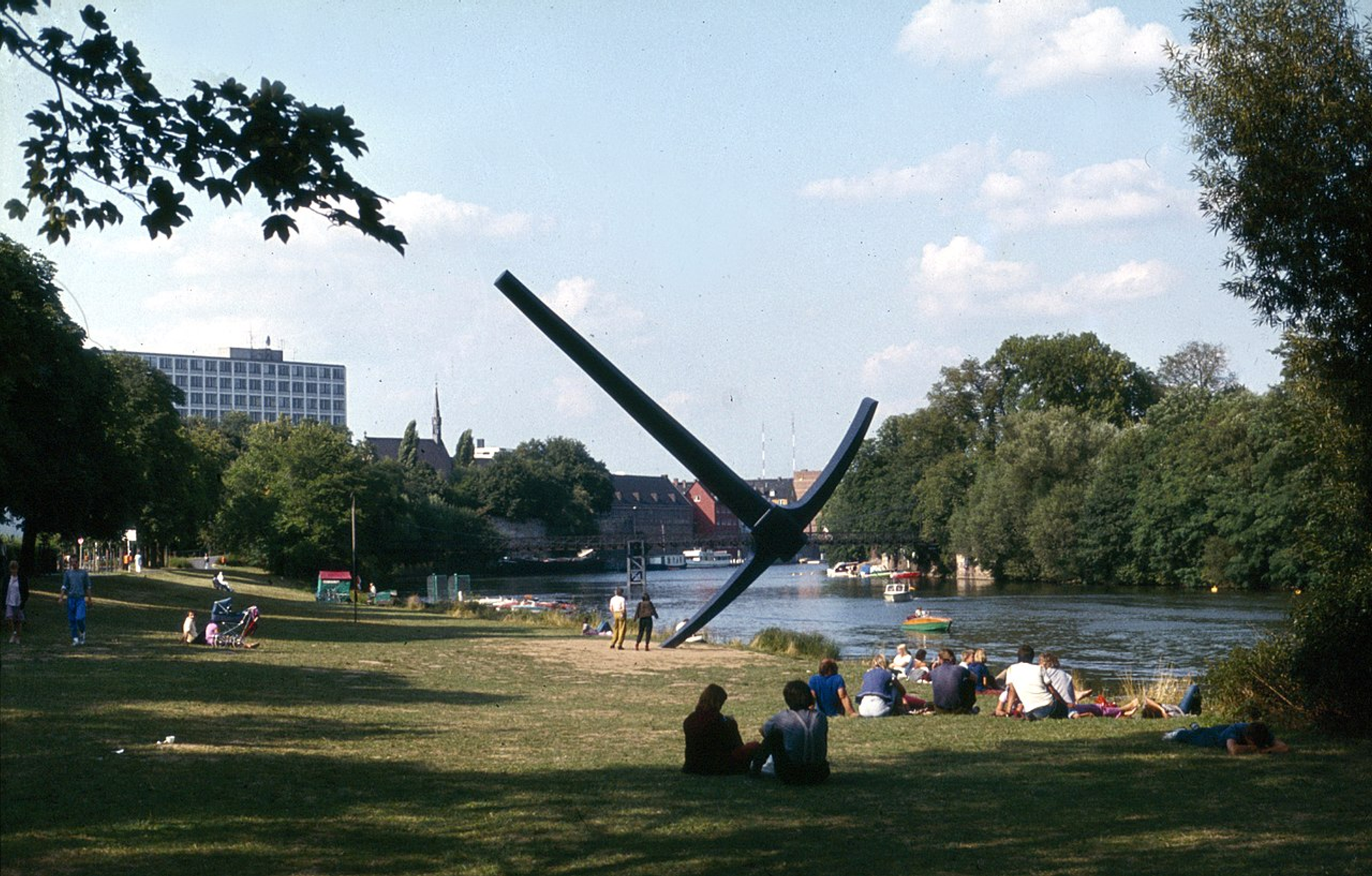 A photograph of one of the outdoor installations carried out for documenta 7. It shows people relaxing in the grass of a park by a lake, with a giant pick axe sculpture by Claes Oldenburg.