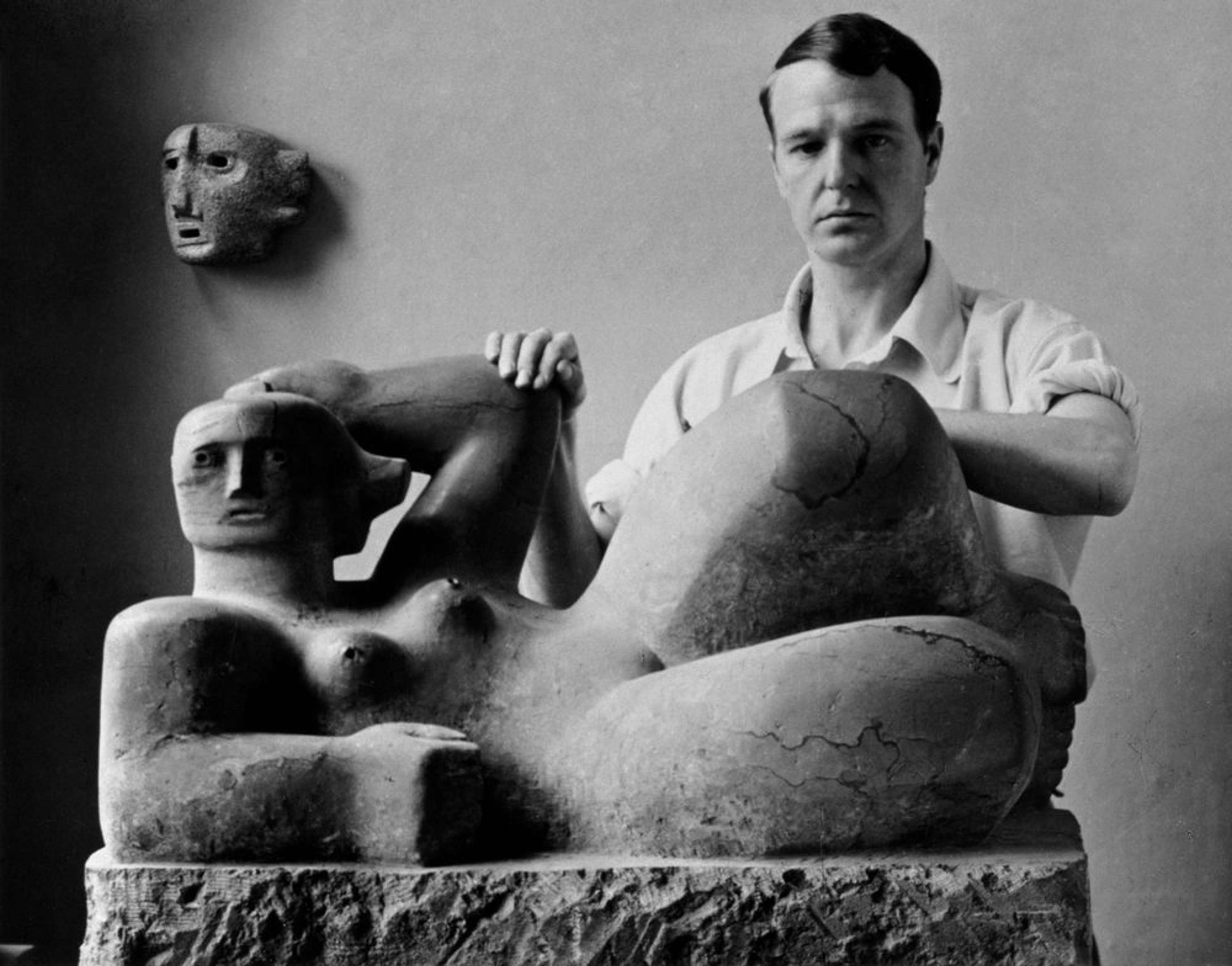 Black-and-white photograph of Henry Moore standing next to a large-scale sculpture. Moore, wearing a white short-sleeve shirt, is positioned beside the abstracted reclining figures sculpture. His hands rest on the raised elbow and knee of the artwork as he gazes directly at the camera.