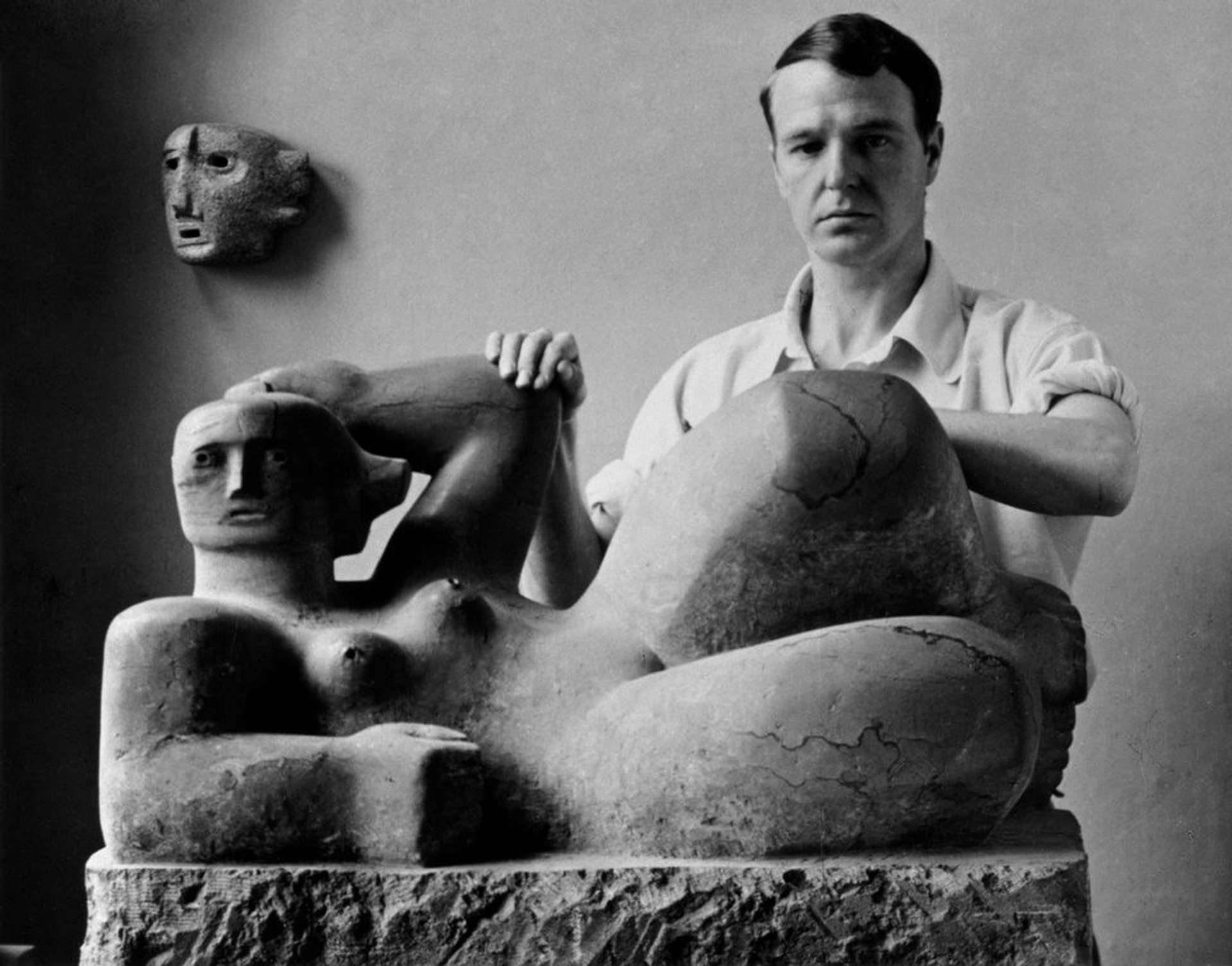 Black-and-white photograph of Henry Moore standing next to a large-scale sculpture. Moore, wearing a white short-sleeve shirt, is positioned beside the abstracted reclining figures sculpture. His hands rest on the raised elbow and knee of the artwork as he gazes directly at the camera.