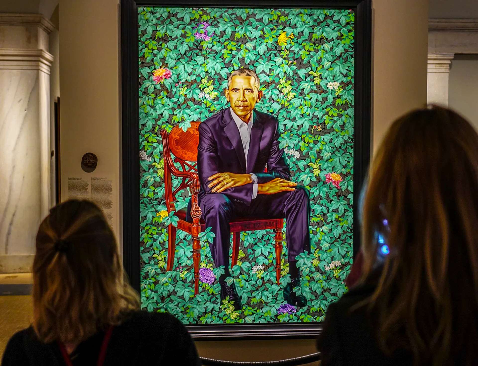 A portrait by Kehinde Wiley of President Barack Obama. He is seated in a chair with his arms folded across his lap wearing a dark suit. He’s placed in front of a brightly coloured background of green foliage.