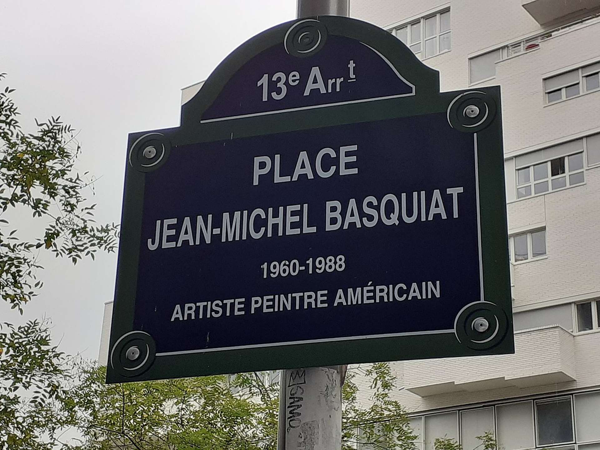 A photograph of the street sign for Place Jean-Michel Basquiat, Paris. Below the sign, on the pole, is a SAMO© tag.