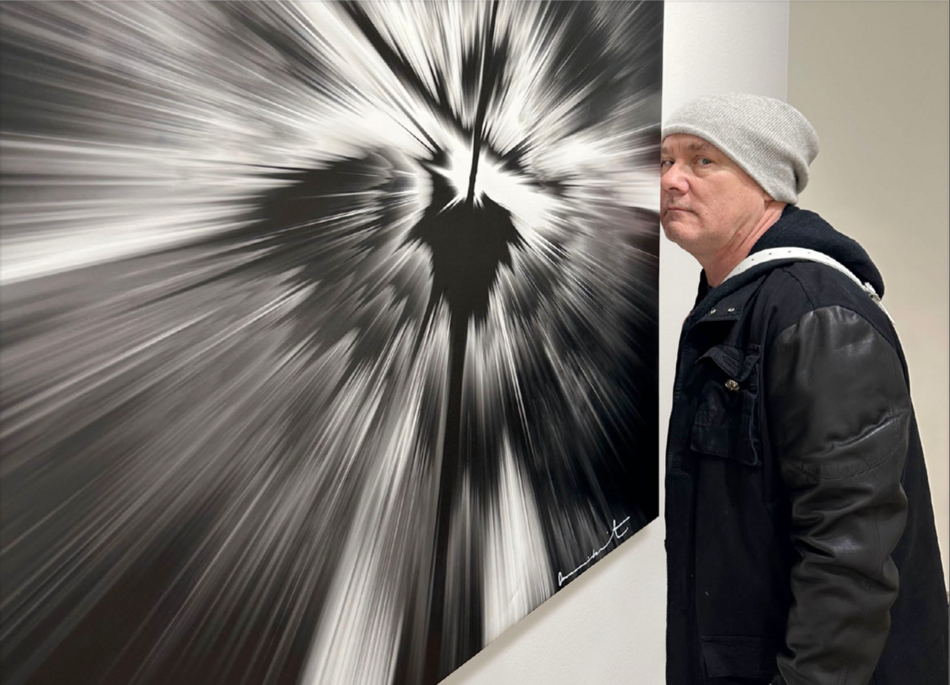Artist Damien Hirst, dressed in a black leather jacket, standing next to one of his Spin Paintings. The printed artwork is in square format, and is composed of black and white, wit the artist's signature in the bottom right corner.