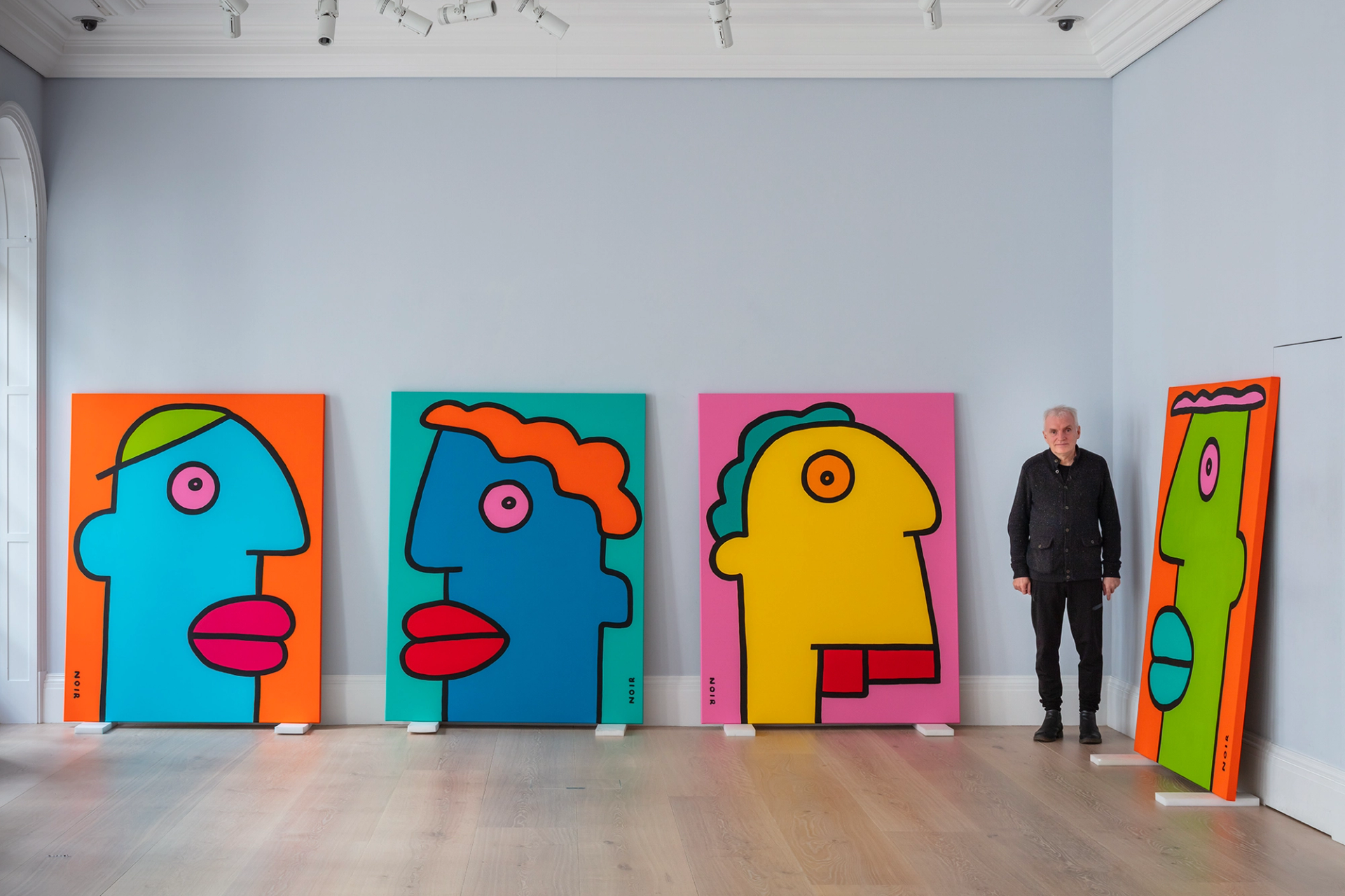 A photograph of Thierry Noir facing the camera and standing in an art gallery surrounded by four large-scale caricature portrait canvases. The canvases are created in vibrant hues and lined up against the walls of the gallery.