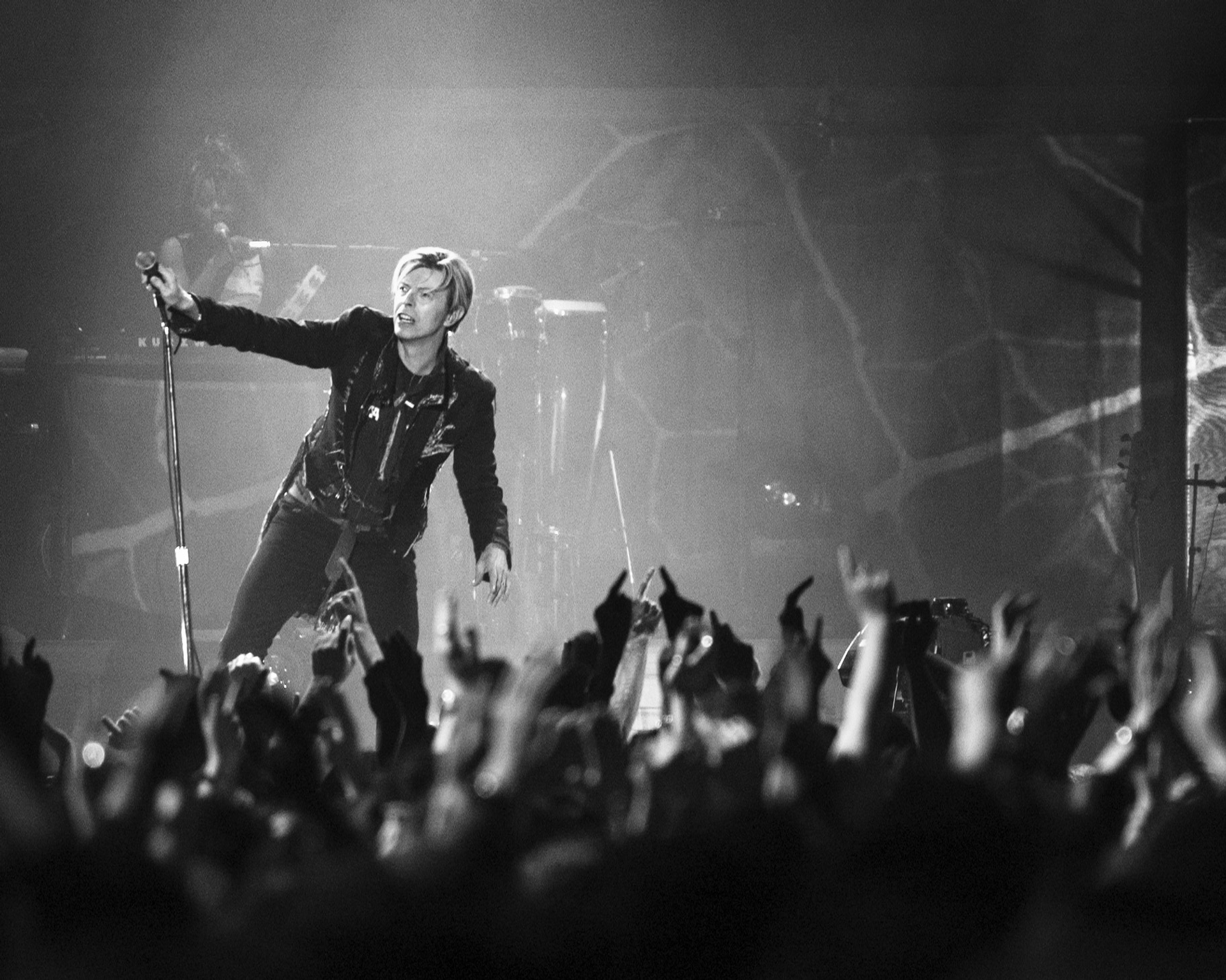 A black-and-white photograph of the musician David Bowie, standing on stage in front of a large crowd. He is wearing black and leaning against the microphone stand.