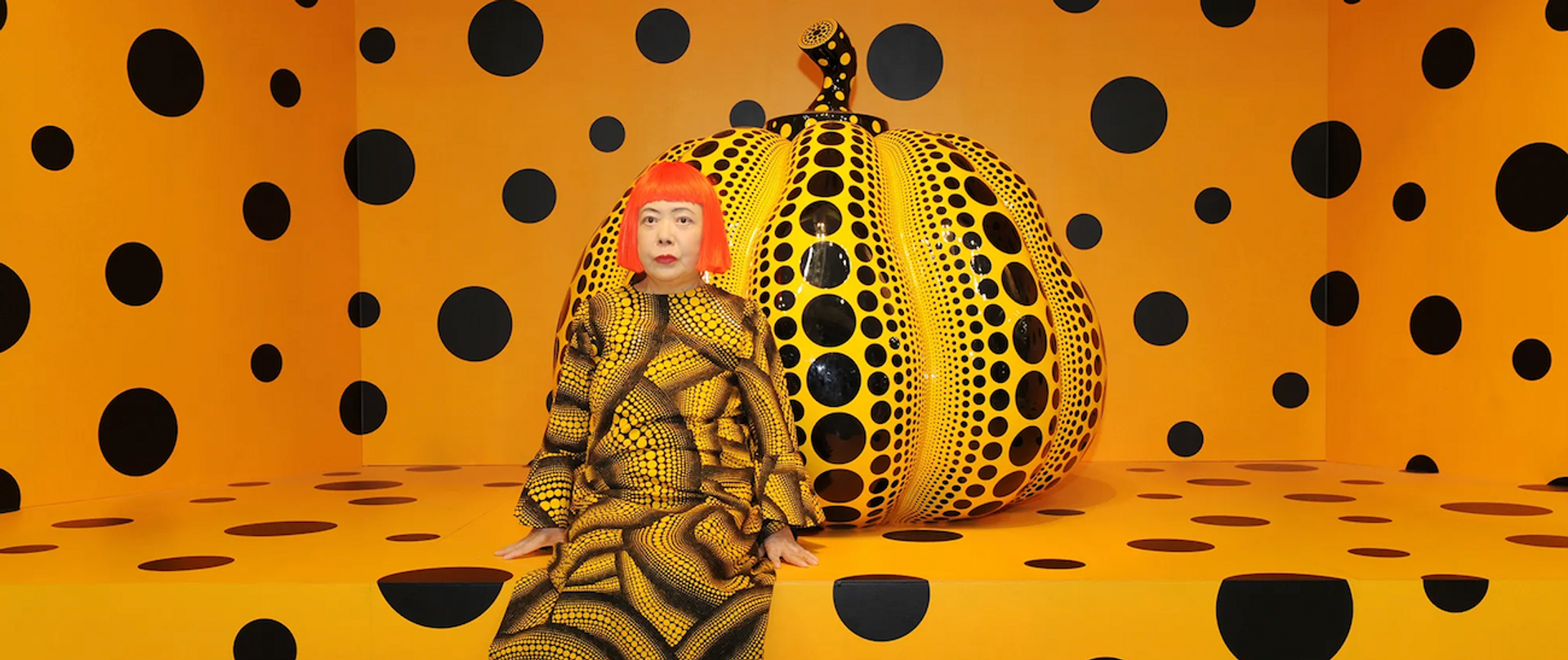 Yayoi Kusama sat in front of a human sized yellow and black pumpkin in an orange room covered in black dots