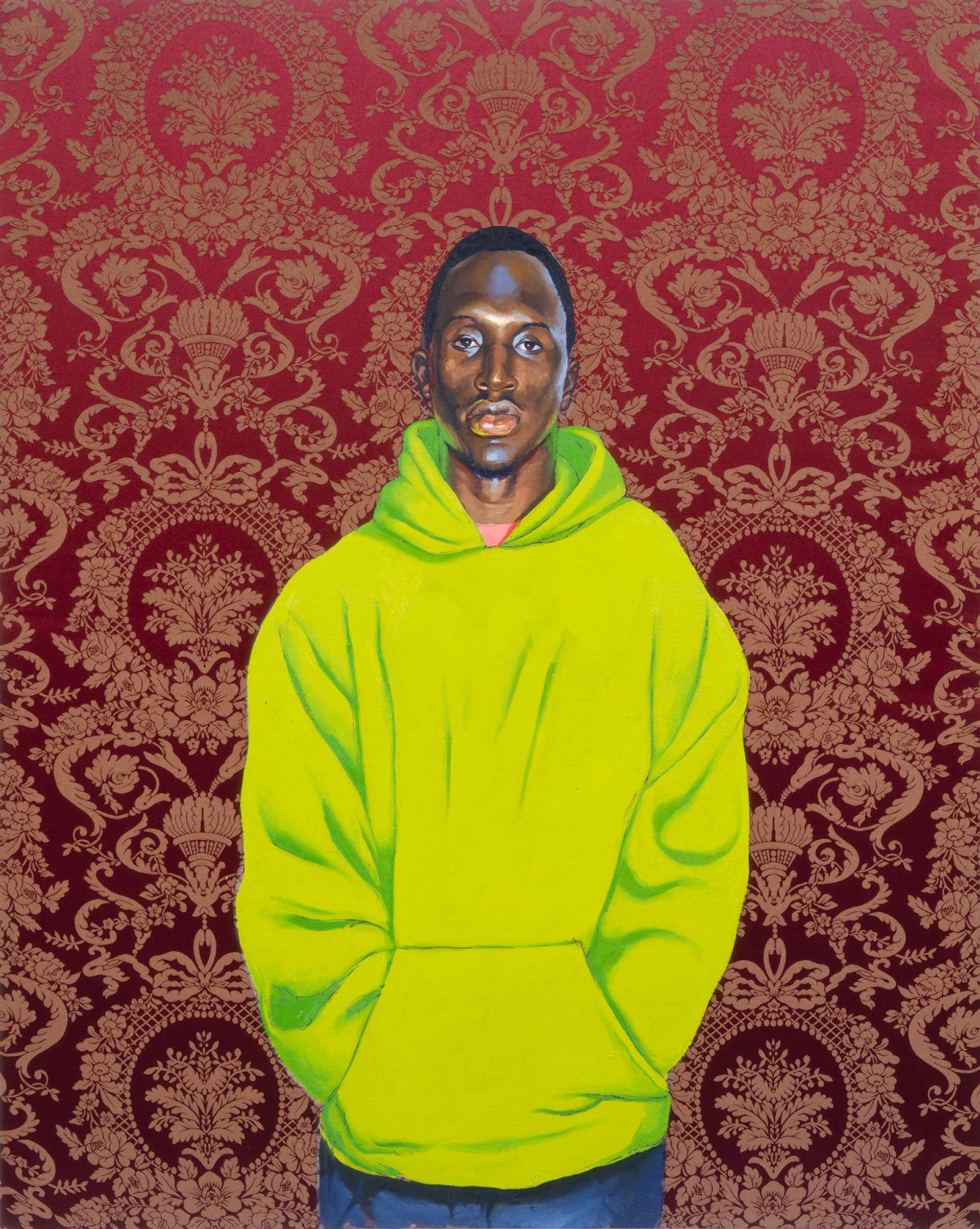A young man looks directly at the viewer; he wears a lime green hoodie and has his hands in his pockets. The brown wallpaper behind him is adorned with an intricate, regal pattern.