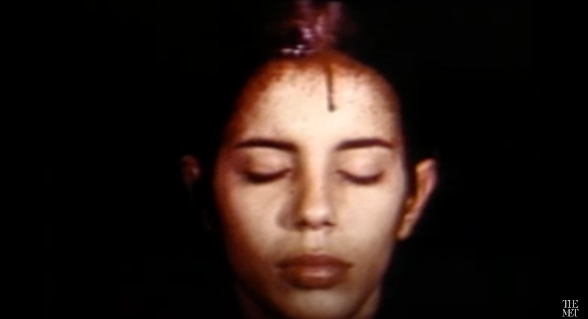 An image of the artist Ana Mendieta from one of her performances. She has her eyes closed as a red substance drips over her forehead.