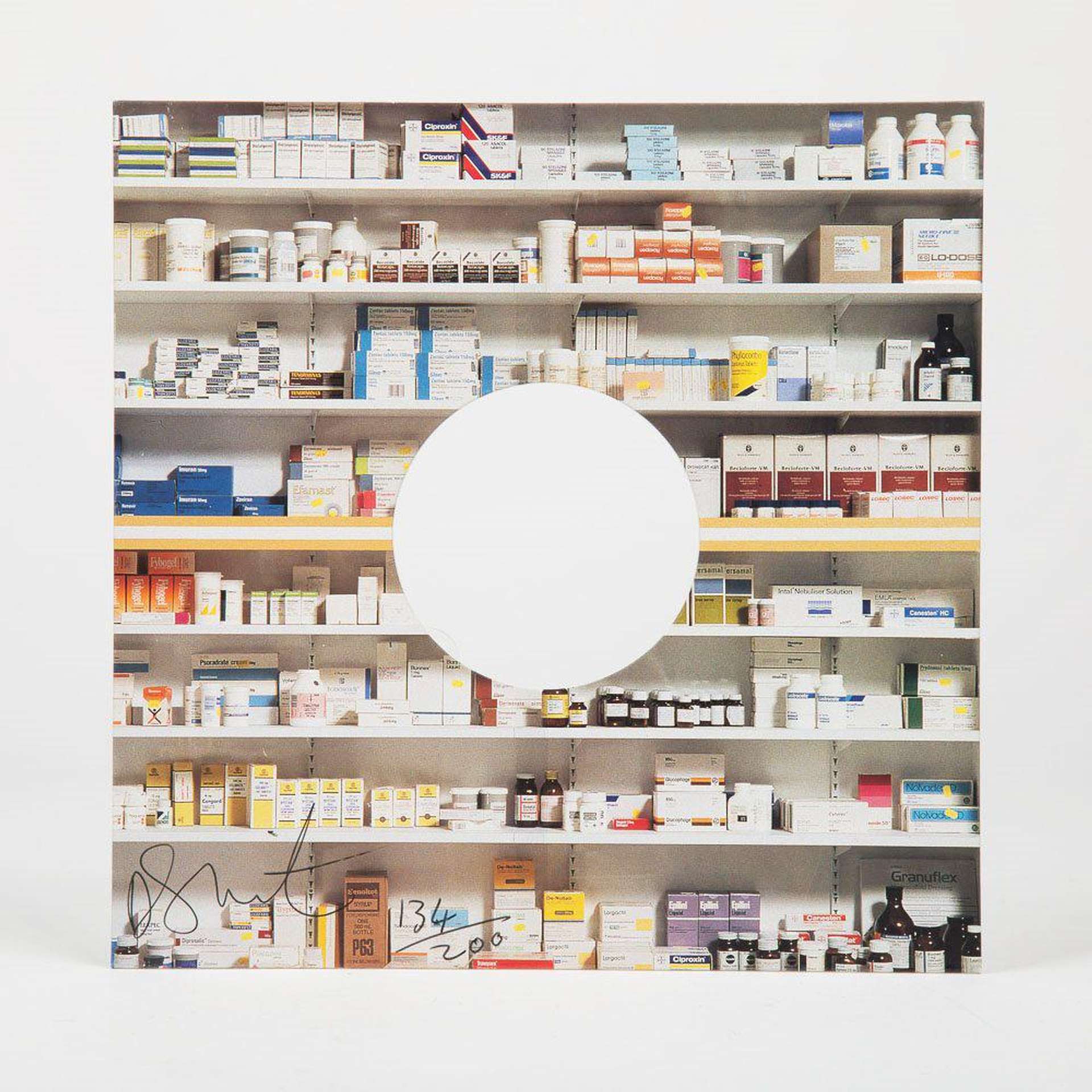 A photograph of Hirst’s Pharmacy, a true to life sized installation of a pharmacy, using materials like shelved cabinets and objects representing pharmaceutical drugs. The print has a white circle in the centre and is framed in white.