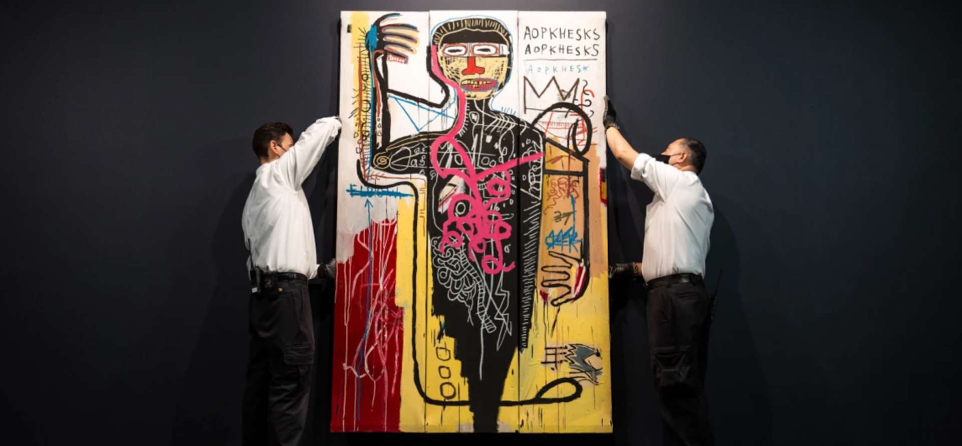 An image of the artwork Versus Medici being carried by two art handlers at Sotheby’s. It shows a figure in black, surrounded by Basquiat’s motifs of scribbling and crowns.