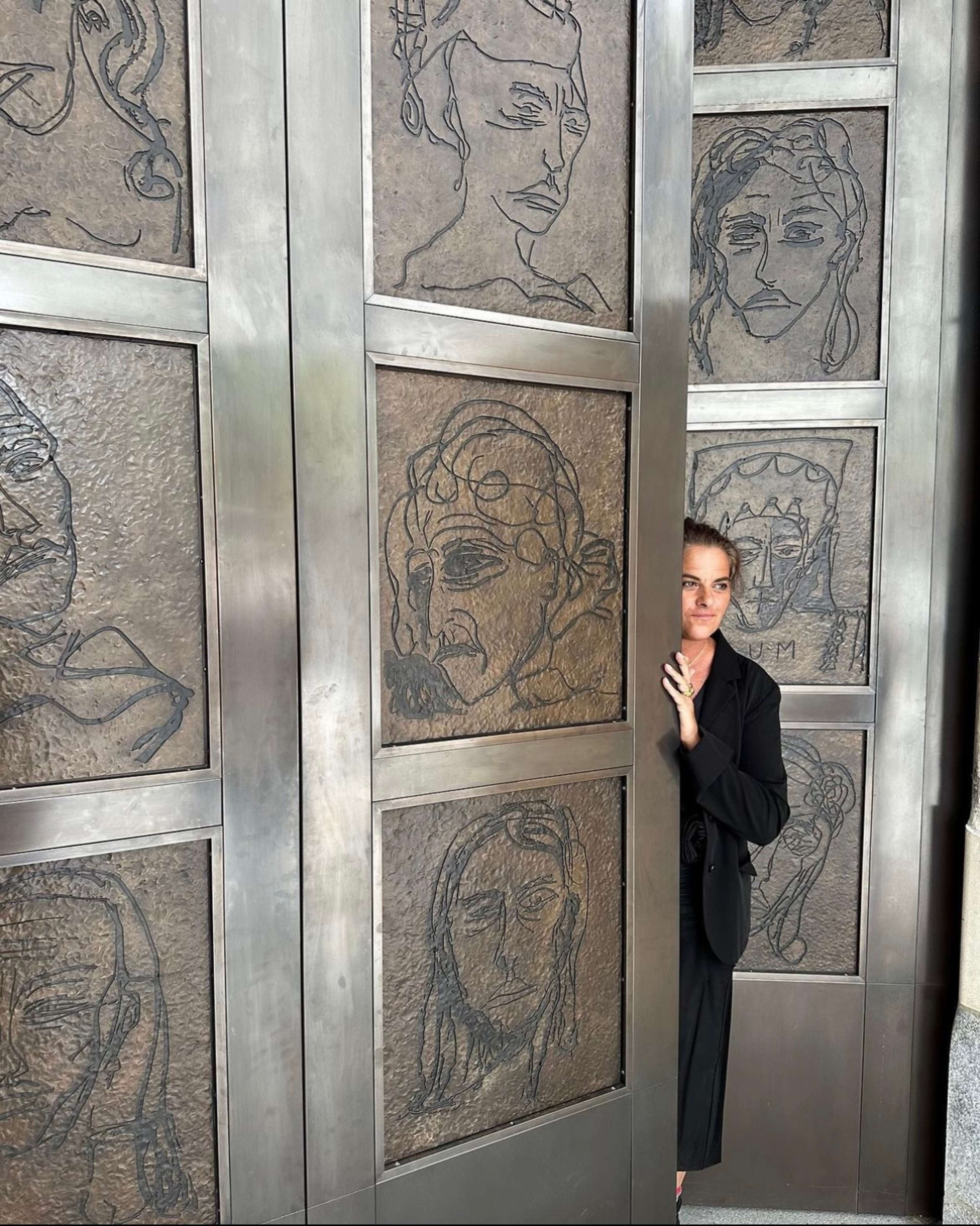 A photograph of artist Tracey Emin, standing in the centre of two doors that she created for the National Portrait Gallery, London. The doors are large and bronze, and feature line drawings of different women. Emin is wearing black and smiling at the camera.