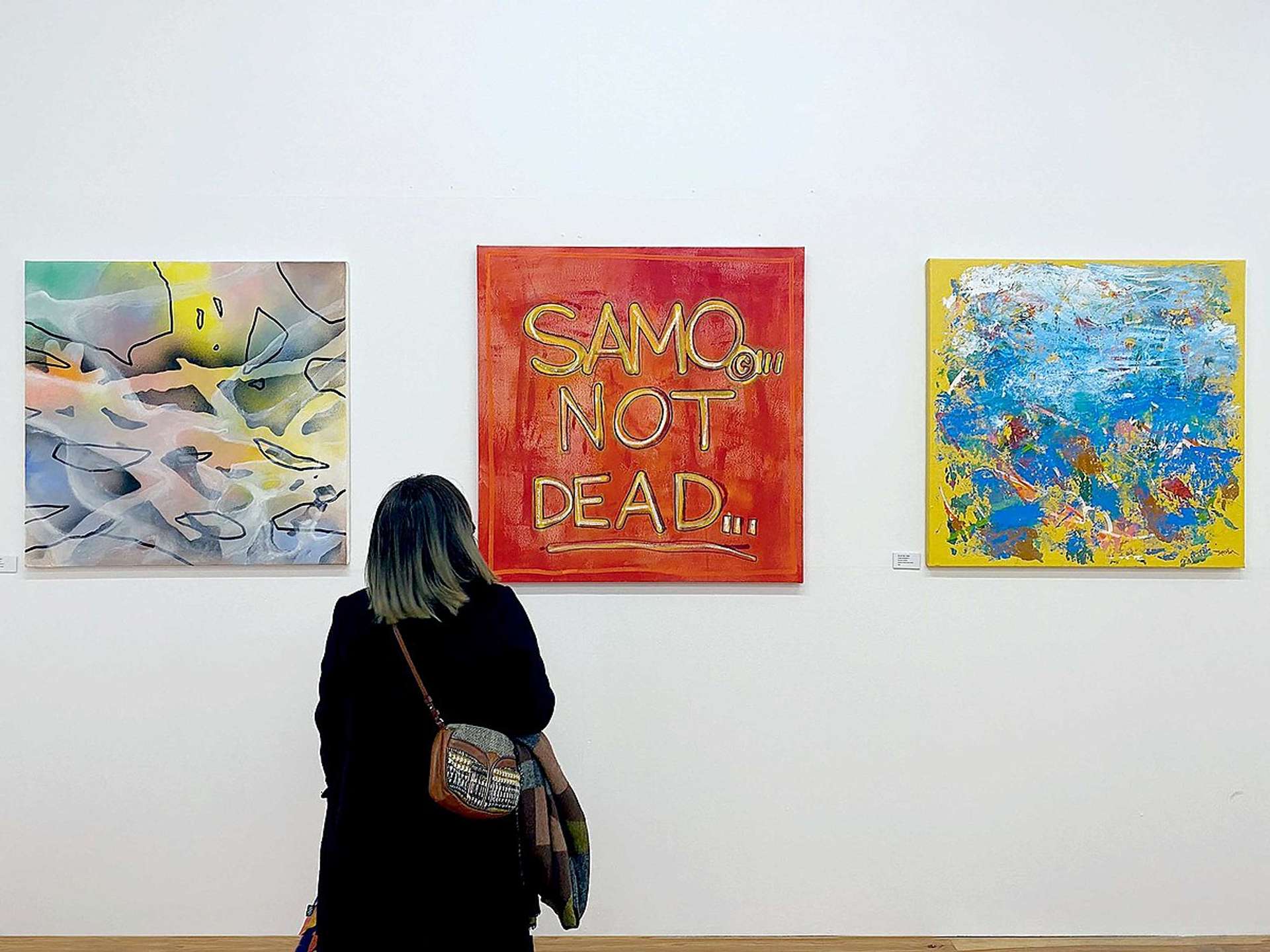 Samo Is Not Dead canvas by Al Diaz. The work is pictured in the centre of a row of three hung canvases, with a woman stood in the foreground, looking at the art.