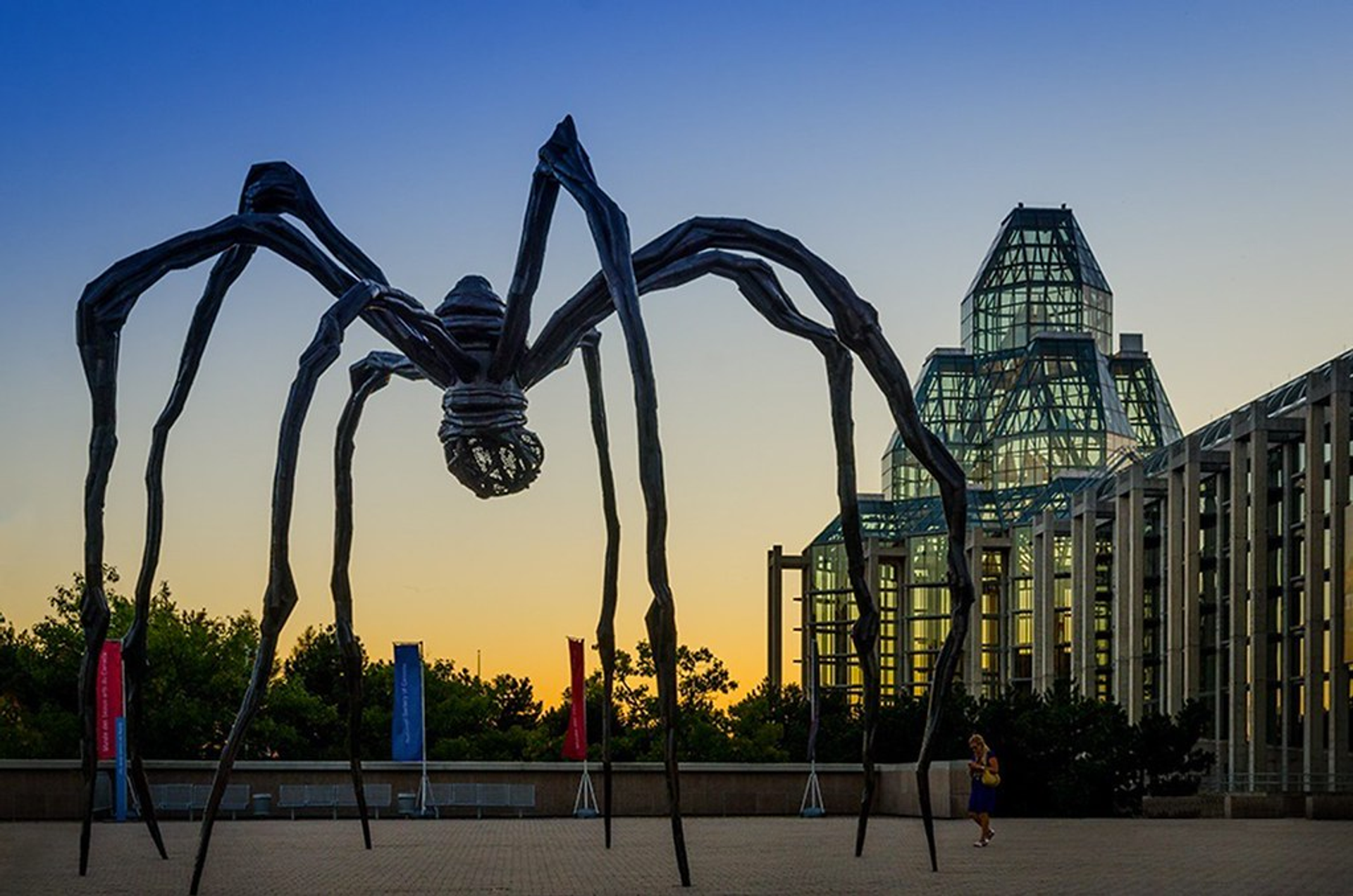 A giant, skinny metal spider sculpture by Louise Bourgeois looms against a sunset background. 