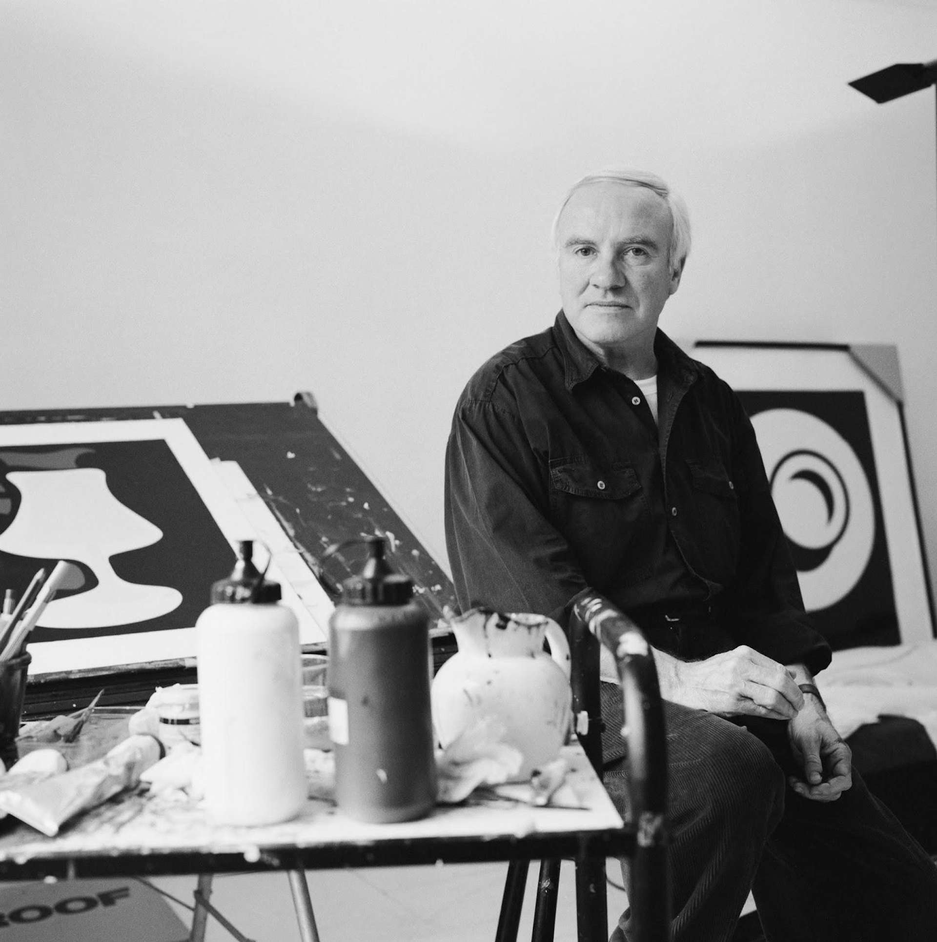 A black and white photographic image of artist Patrick Caulfield, standing in front of several of his works in his studio.