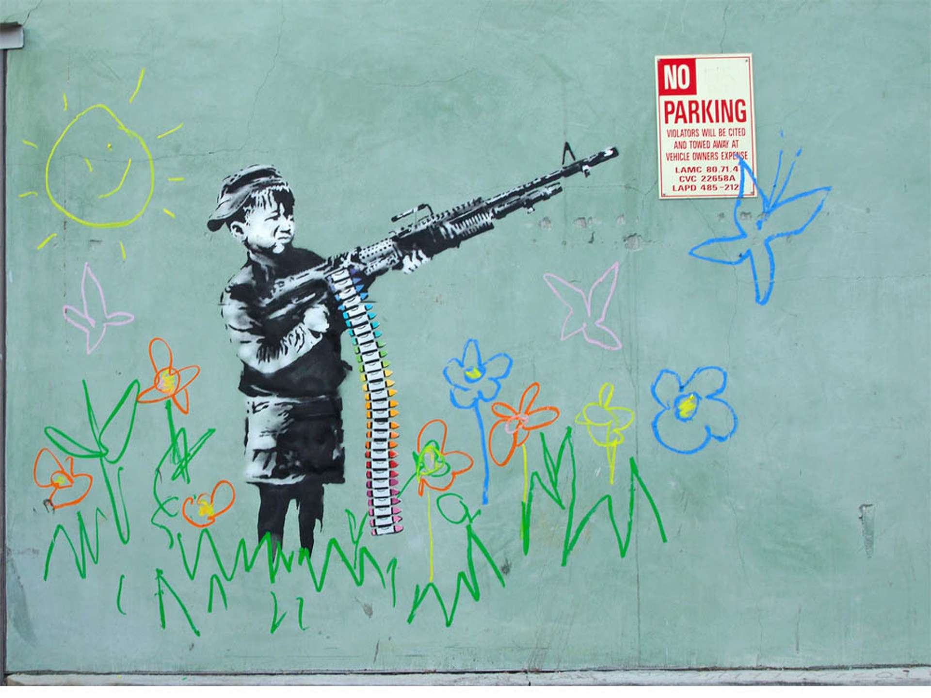 An image of the mural Crayon Boy by Banksy. It shows a monochrome young boy holding a machine gun, but its cartridges are made of crayons. Around him, are child-like depictions of nature such as flowers and a smiling sun.