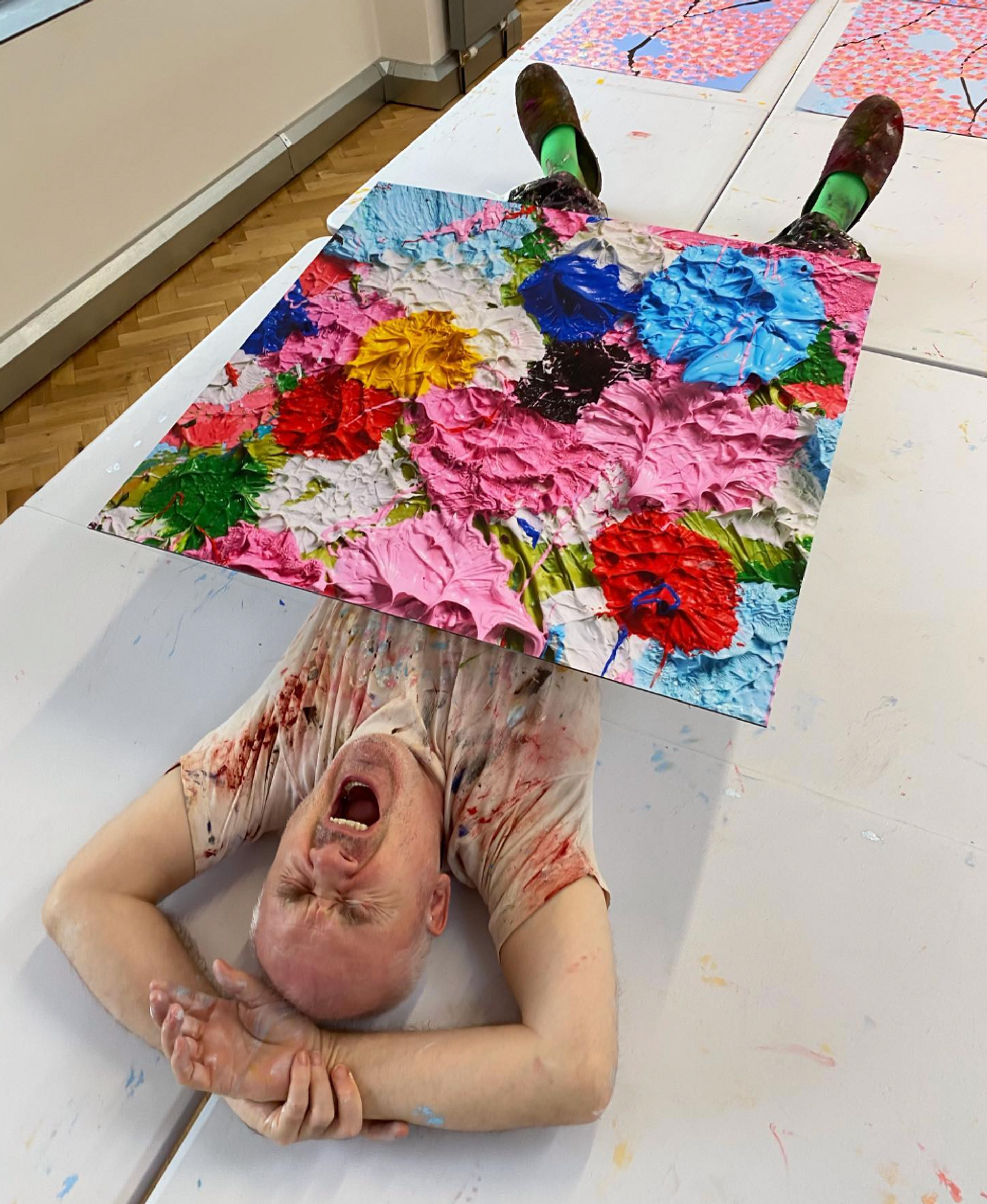 An image of the artist Damien Hirst yawning, while the large size of one of his works from the Fruitful And Forever series lies on his stomach.