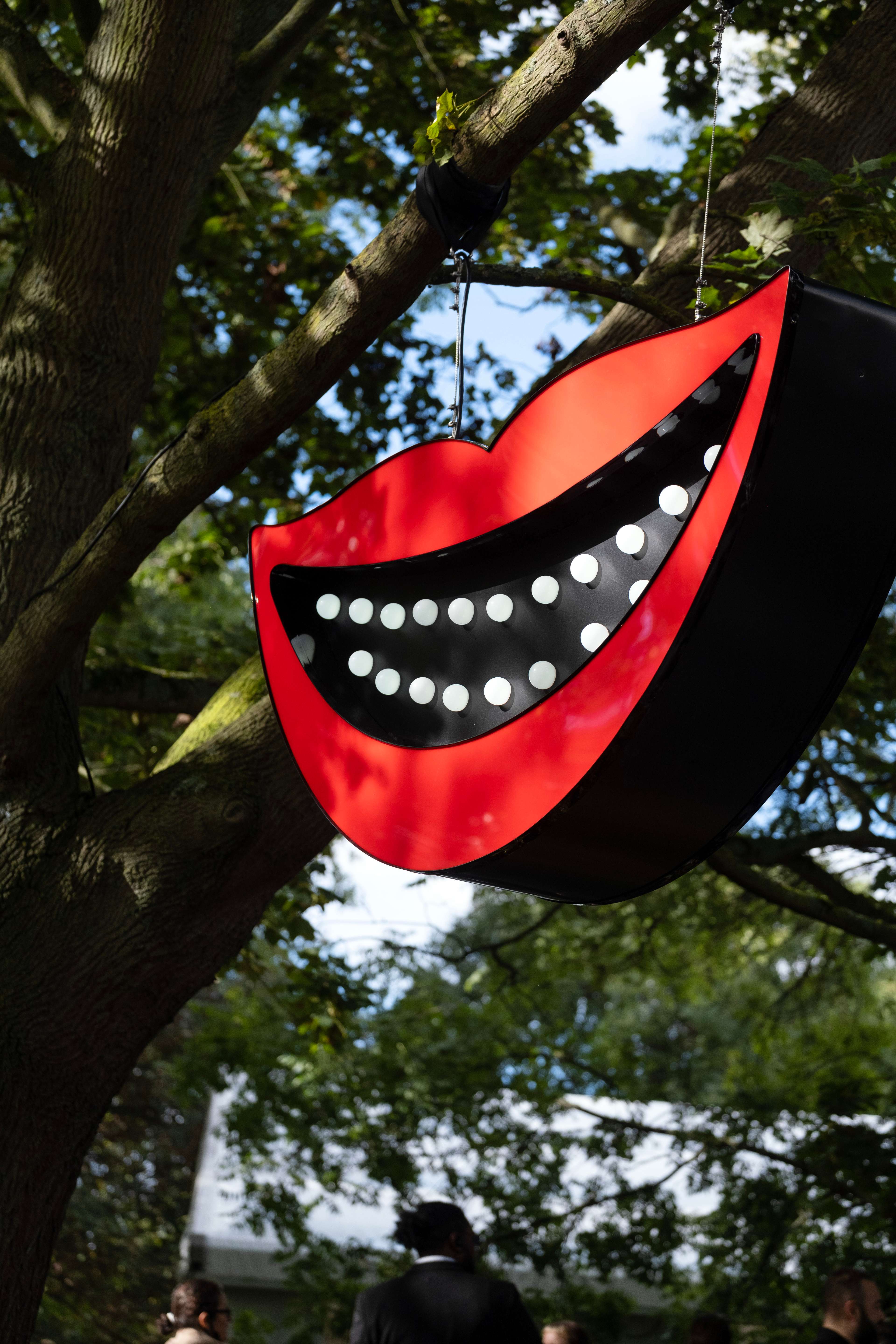Large-scale artwork: Smiling mouth with white lights as teeth, installed in a tree at Frieze Art Fair, London 2023.