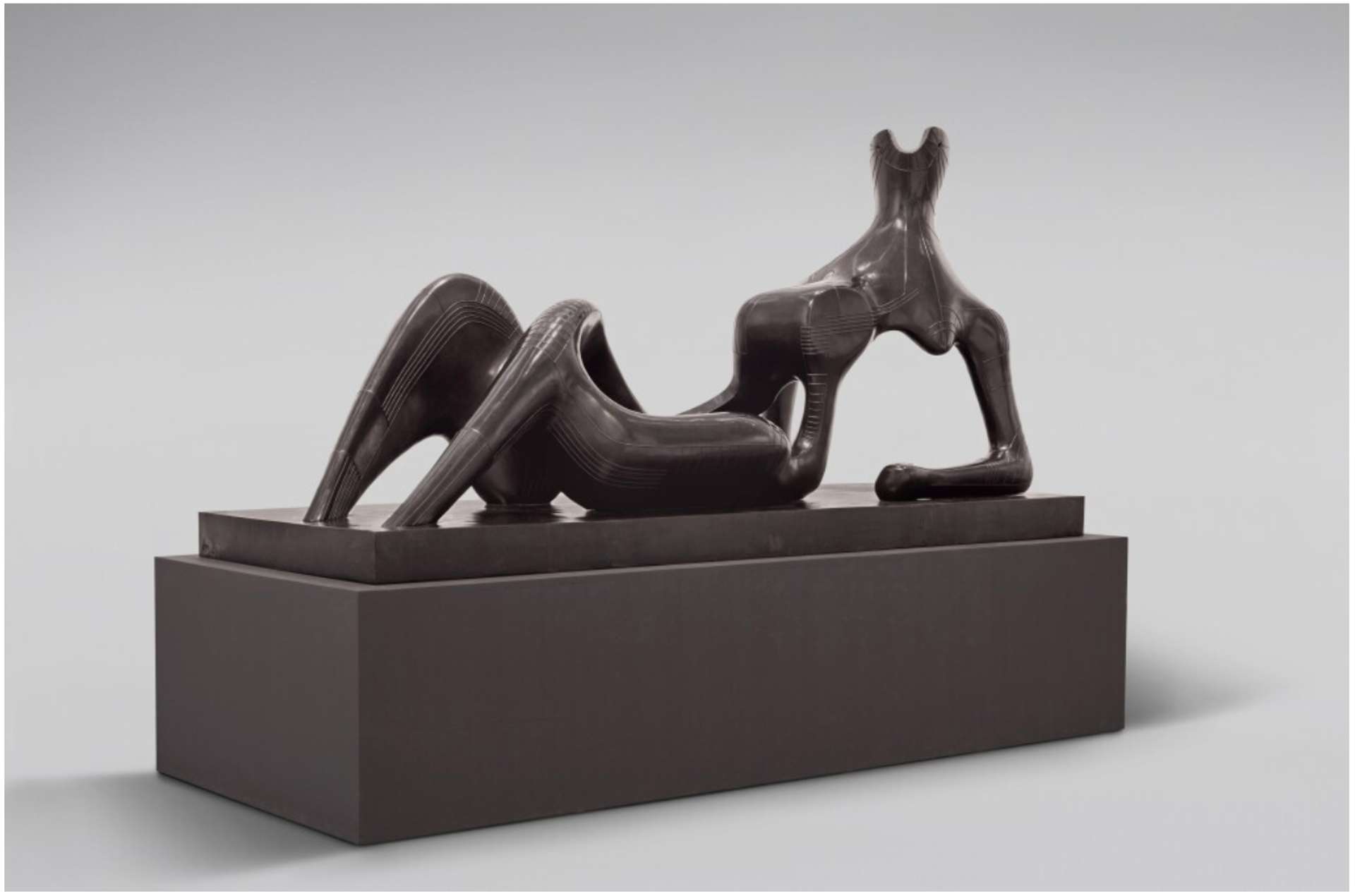 A large-scale bronze sculpture of a biomorphic reclining figure. The sculpture is positioned on a plinth, elevated on a raised platform. The figure's feet are not clearly defined, seamlessly merging into the plinth. The figure rests on a bent elbow, creating a substantial void between the elbow and the upper body representation. The sculpture skillfully balances mass and empty space.