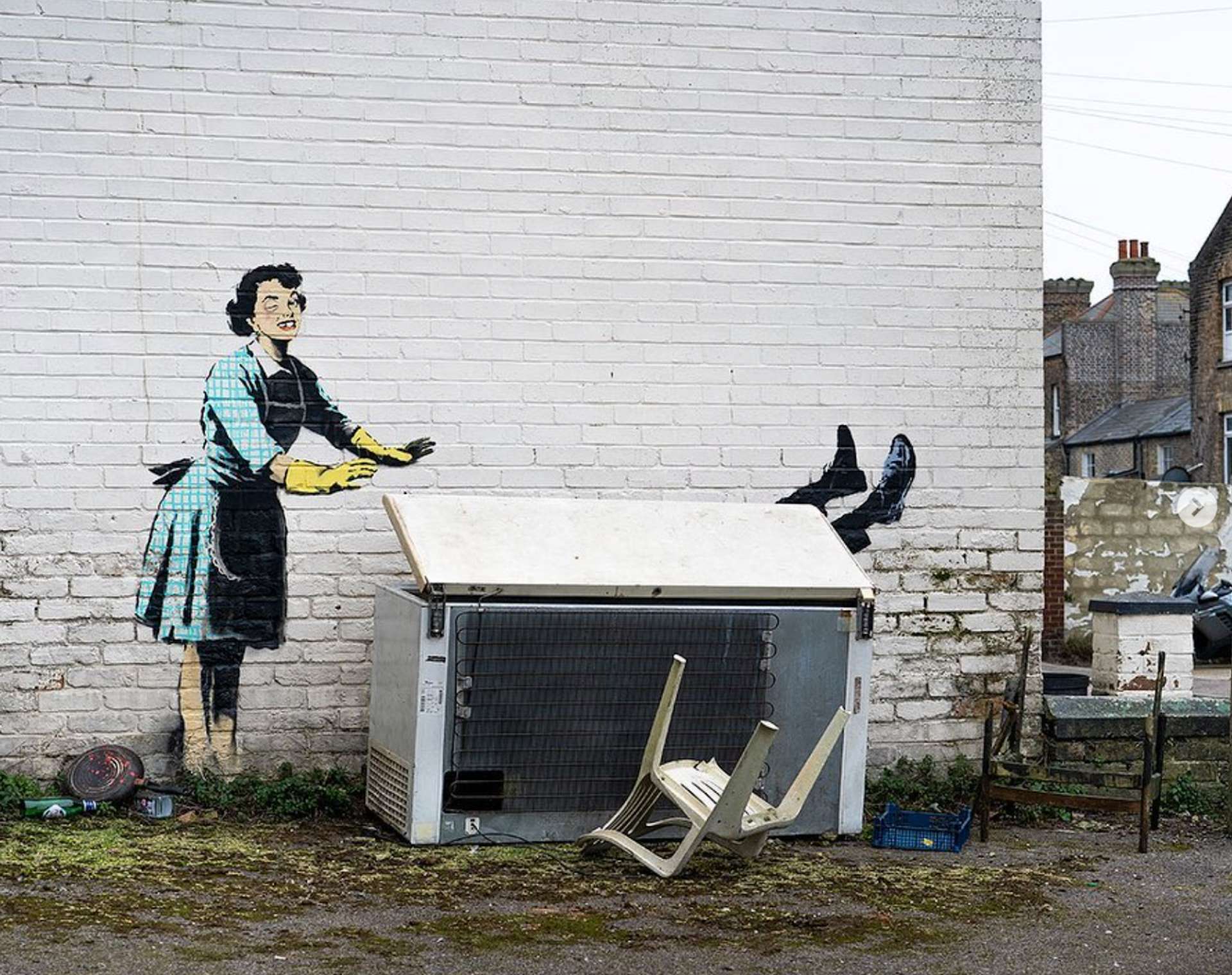This image shows a graffiti of woman, wearing bright yellow dish washing gloves, seemingly closing the lid on a real discarded freezer, from which a pair of feet in male shoes emerges. The woman has a black eye and a swollen cheek, and is missing a tooth.