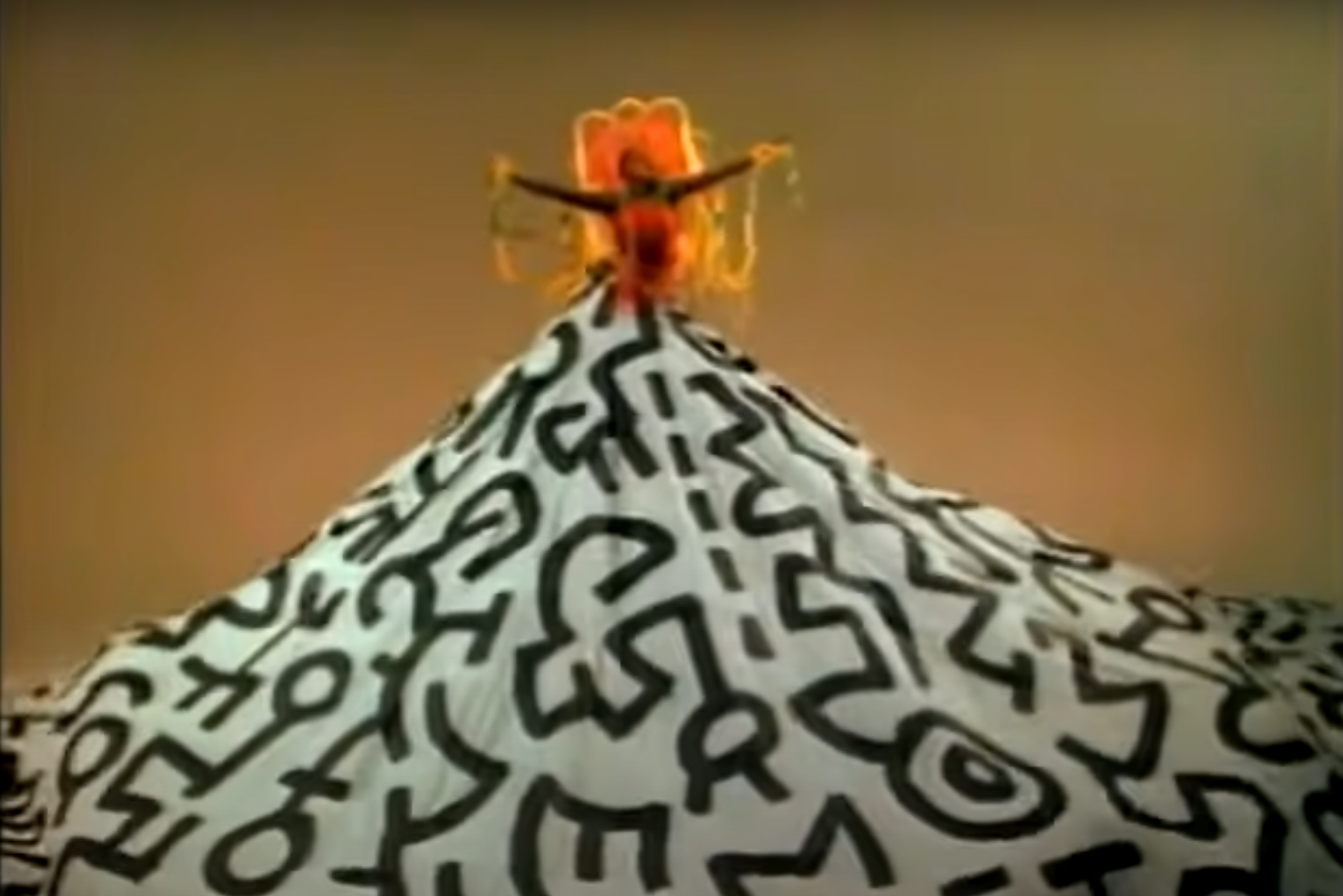 A screenshot of artist Grace Jones, raised up high with a giant skirt decorated with monochromatic Keith Haring motifs.