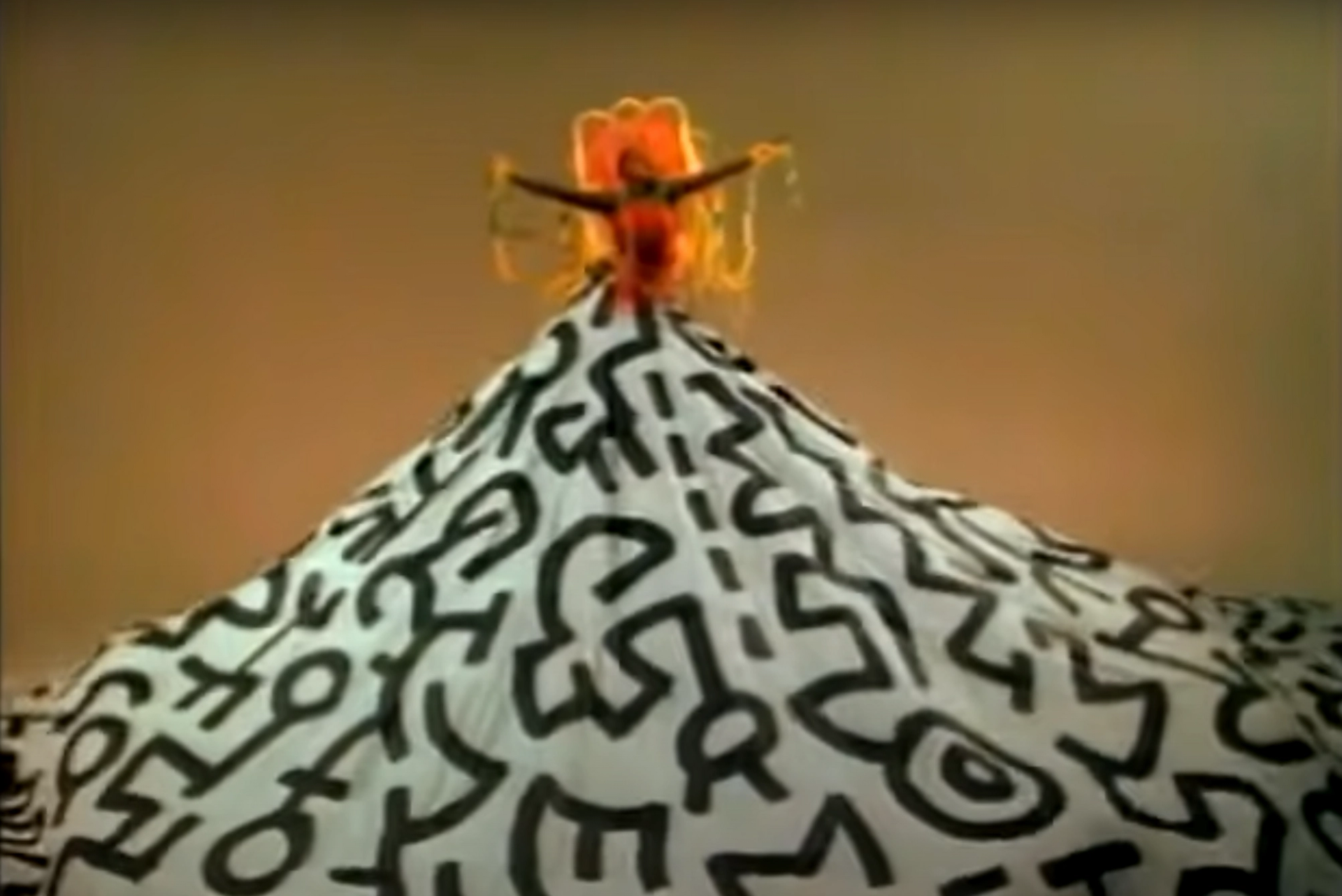 A screenshot of artist Grace Jones, raised up high with a giant skirt decorated with monochromatic Keith Haring motifs.