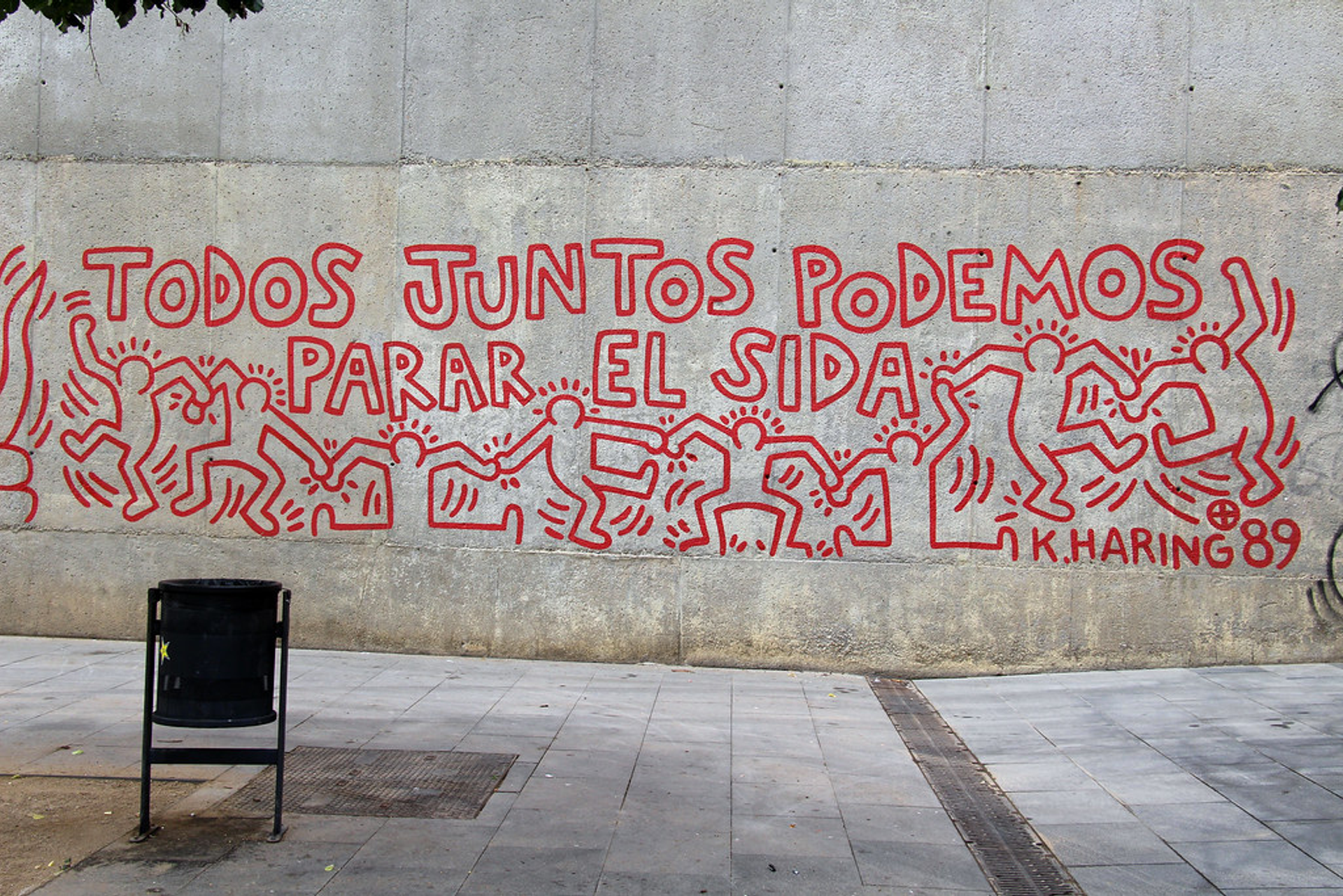 An image of a mural by Keith Haring in Barcelona, Spain. Done in red, it shows his signature interlaced dancing men, around a line of text that says "Together we can stop AIDS" in Spanish.