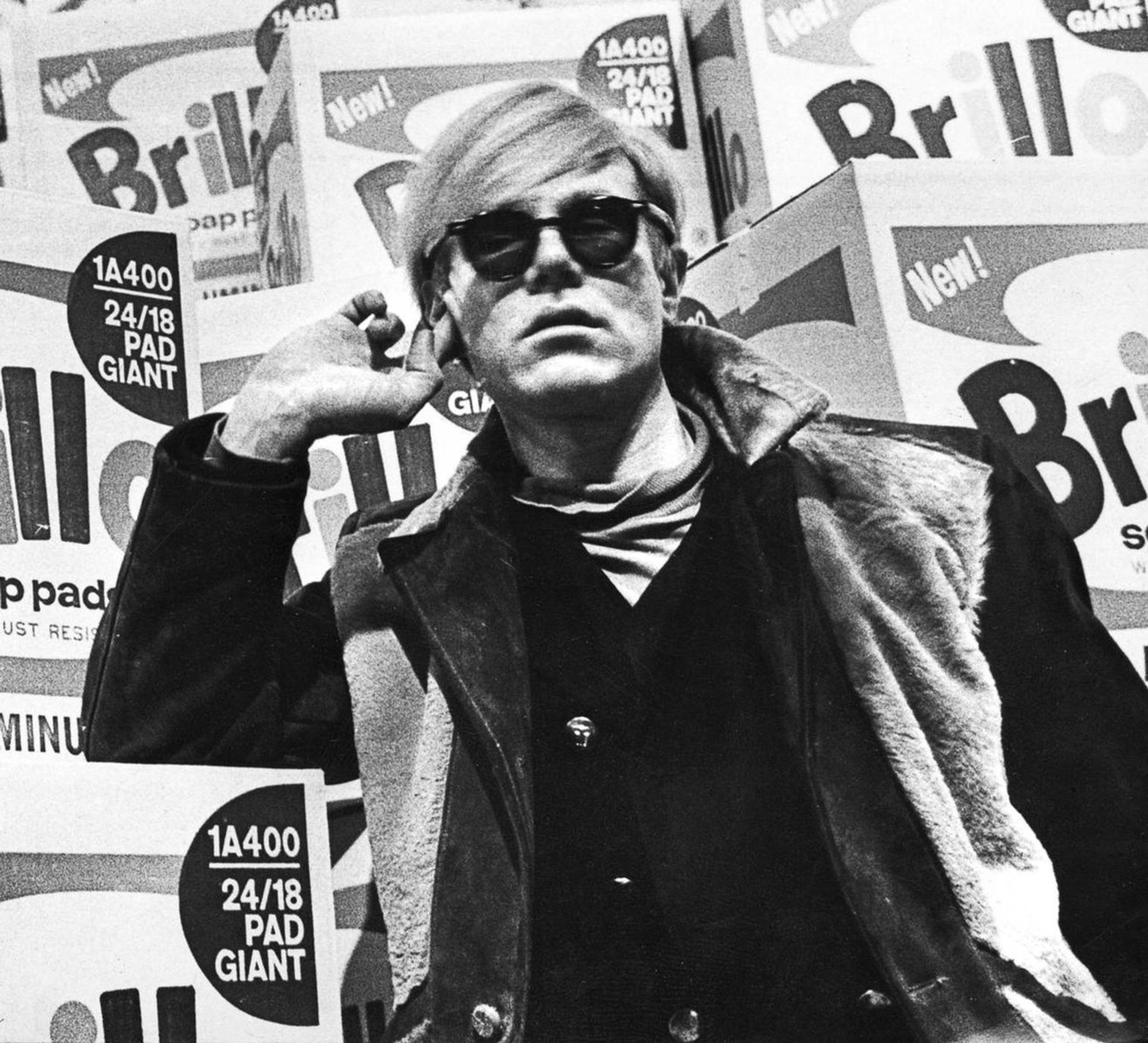 A monochrome image of the artist Andy Warhol standing in front of several of his Brillo boxes. Warhol is wearing a furry gilet and sunglasses.
