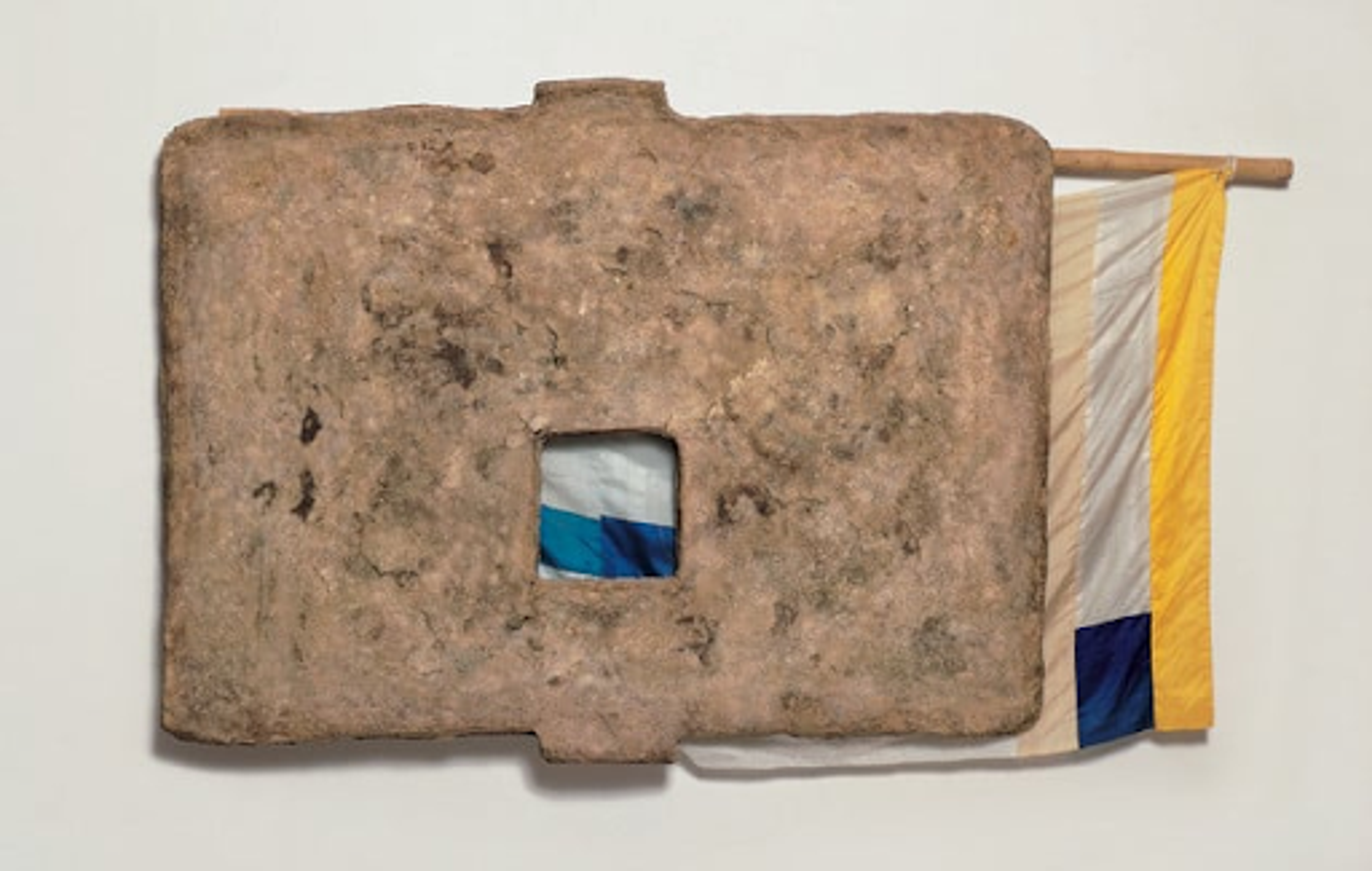 Robert Rauschnberg’s Capitol (Unions). An artwork including a yellow, white, and blue multi-patterned fabric attached to a bamboo stick behind a surface made of “rag-mud”.