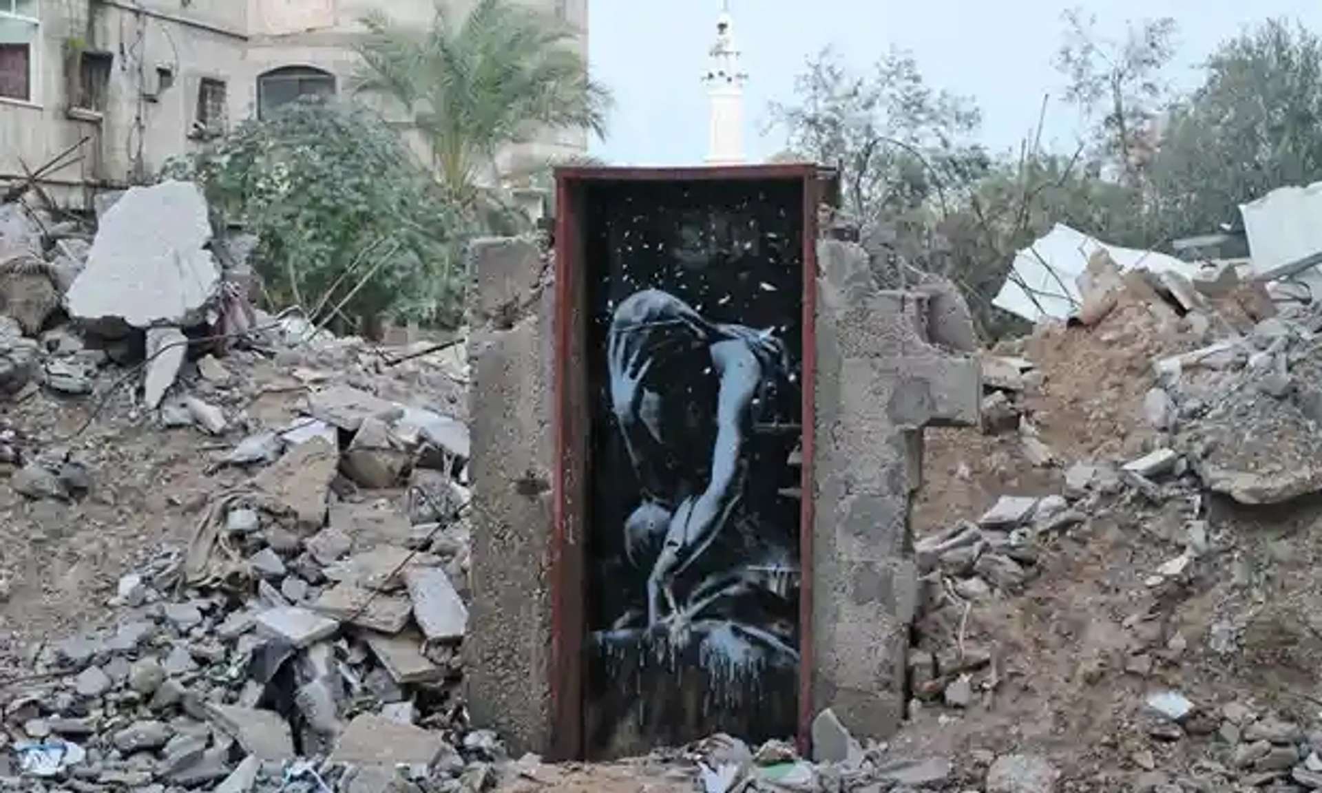 An image of a mural by Banksy amidst the rubble in Gaza. It shows a bent over man, with his head in his hands, depicted in monochrome.