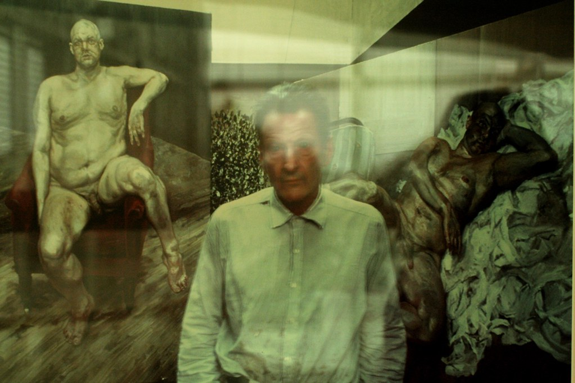 A photograph of the artist Lucian Freud standing in front of two portraits of Leigh Bowery in the nude.