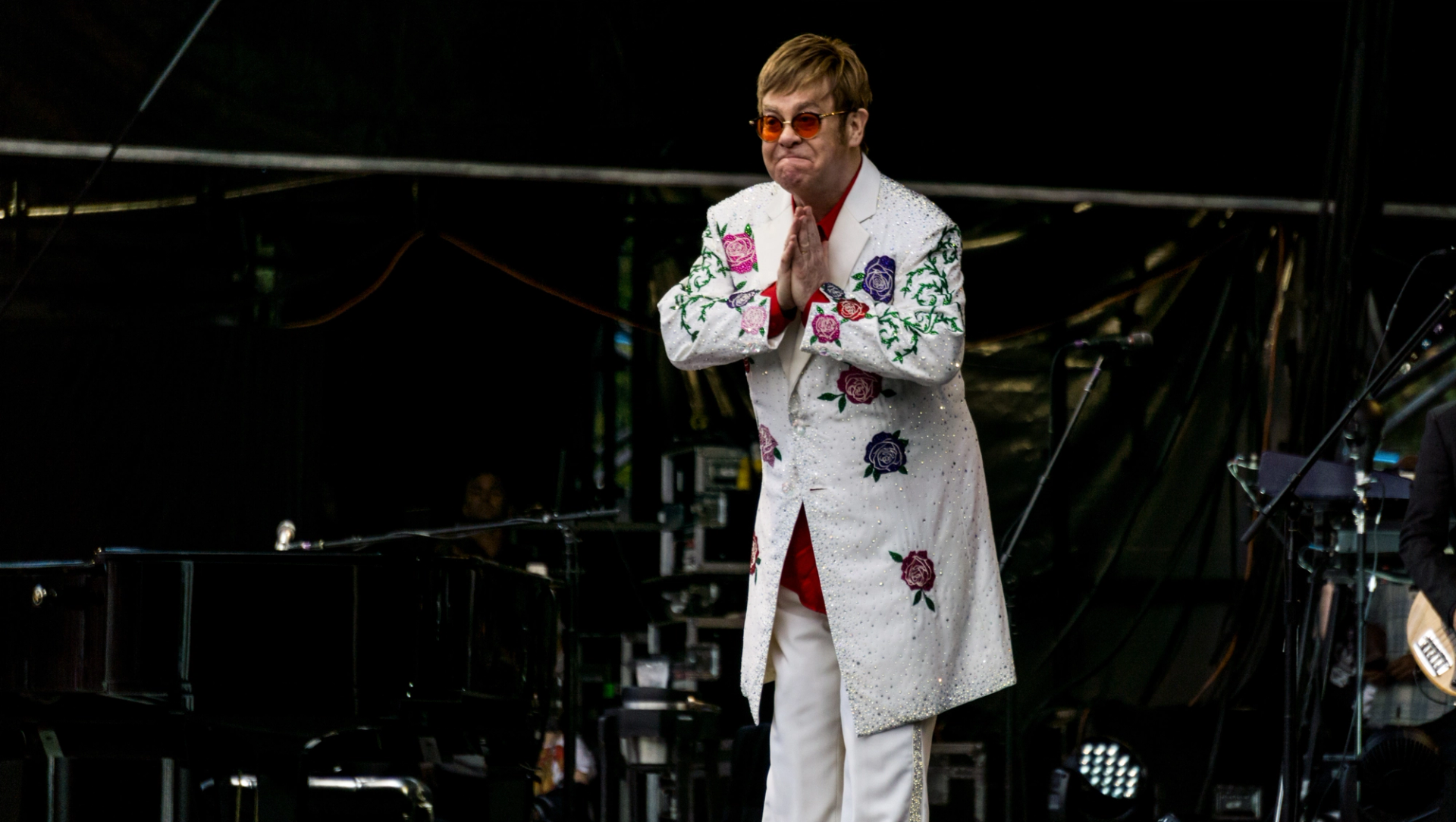 This photograph shows artist Elton John on stage, wearing a white sparkly embroidered suit and his signature colourful sunglasses.