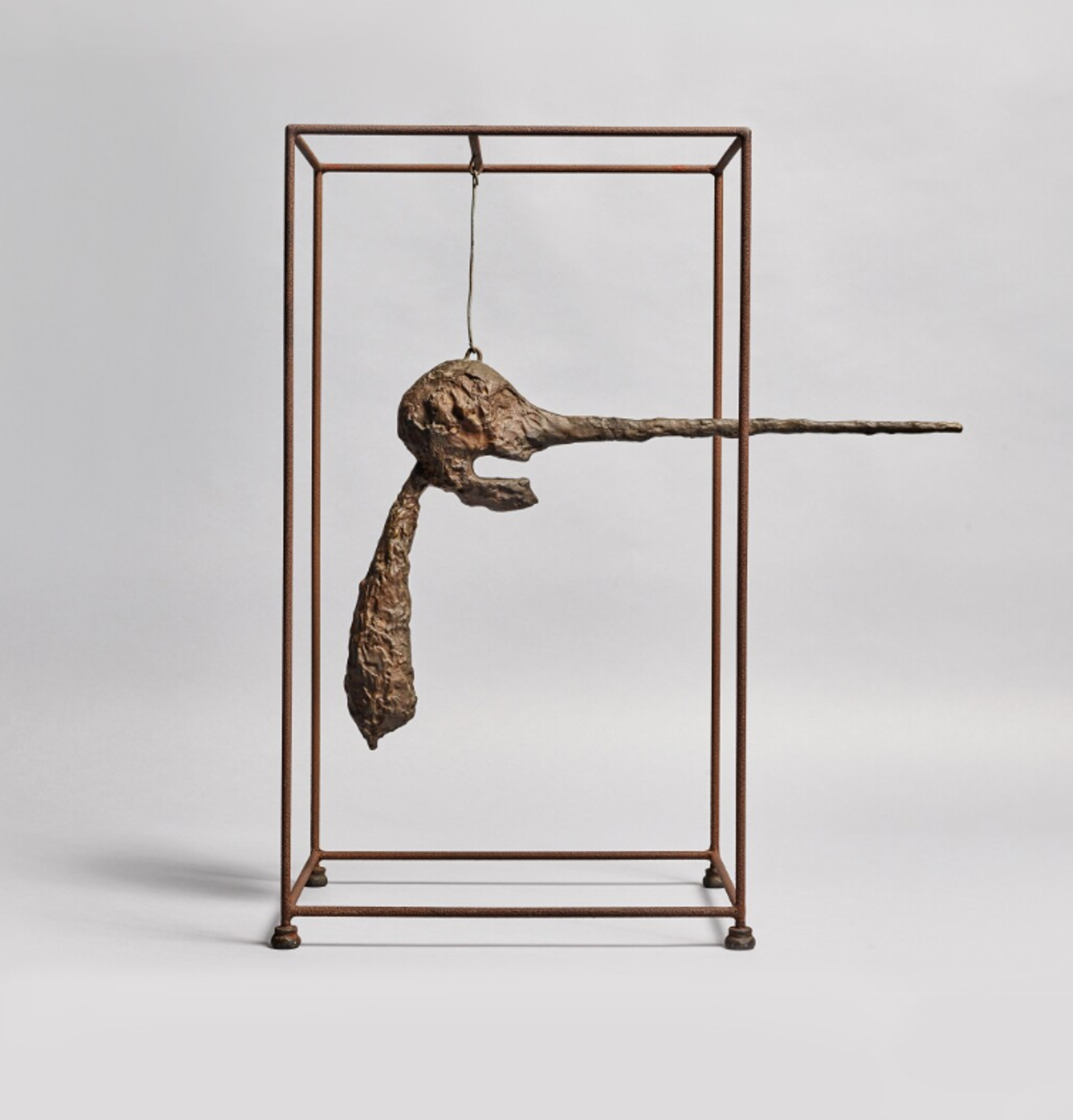 Bronze sculpture featuring a cage-like rectangular frame housing a suspended, elongated head form with a dramatically protruding, needle-like nose. The end of the nose extends beyond the confines of the cage.
