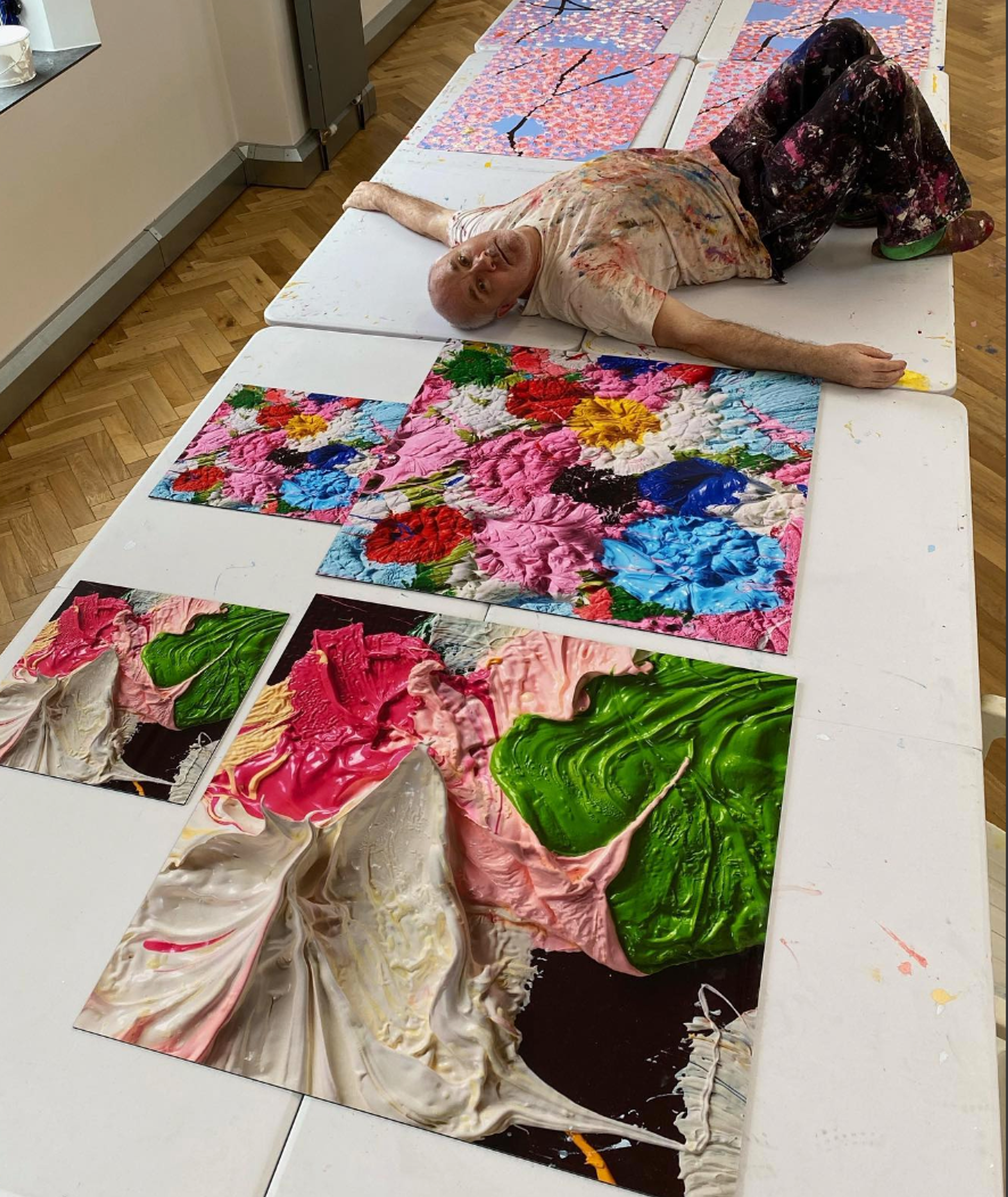 An image of the artist Damien Hirst lying alongside his works from the series Fruitful And Forever. He is shown wearing a paint-splattered outfit.