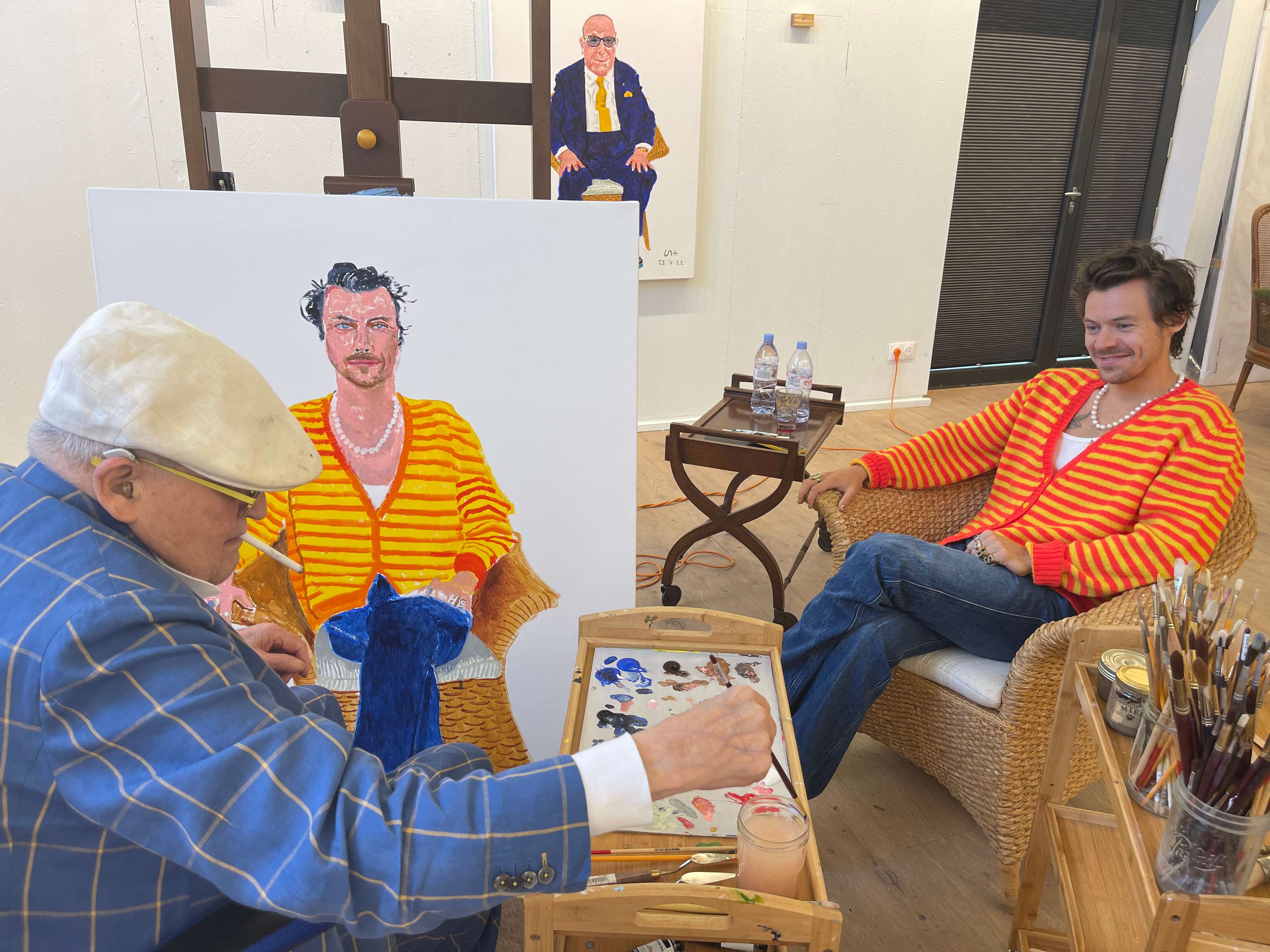 Une photographie montrant l'artiste David Hockney (à gauche) illustrant le portrait de la pop star Harry Styles (à droite), lequel se situe entre les deux.