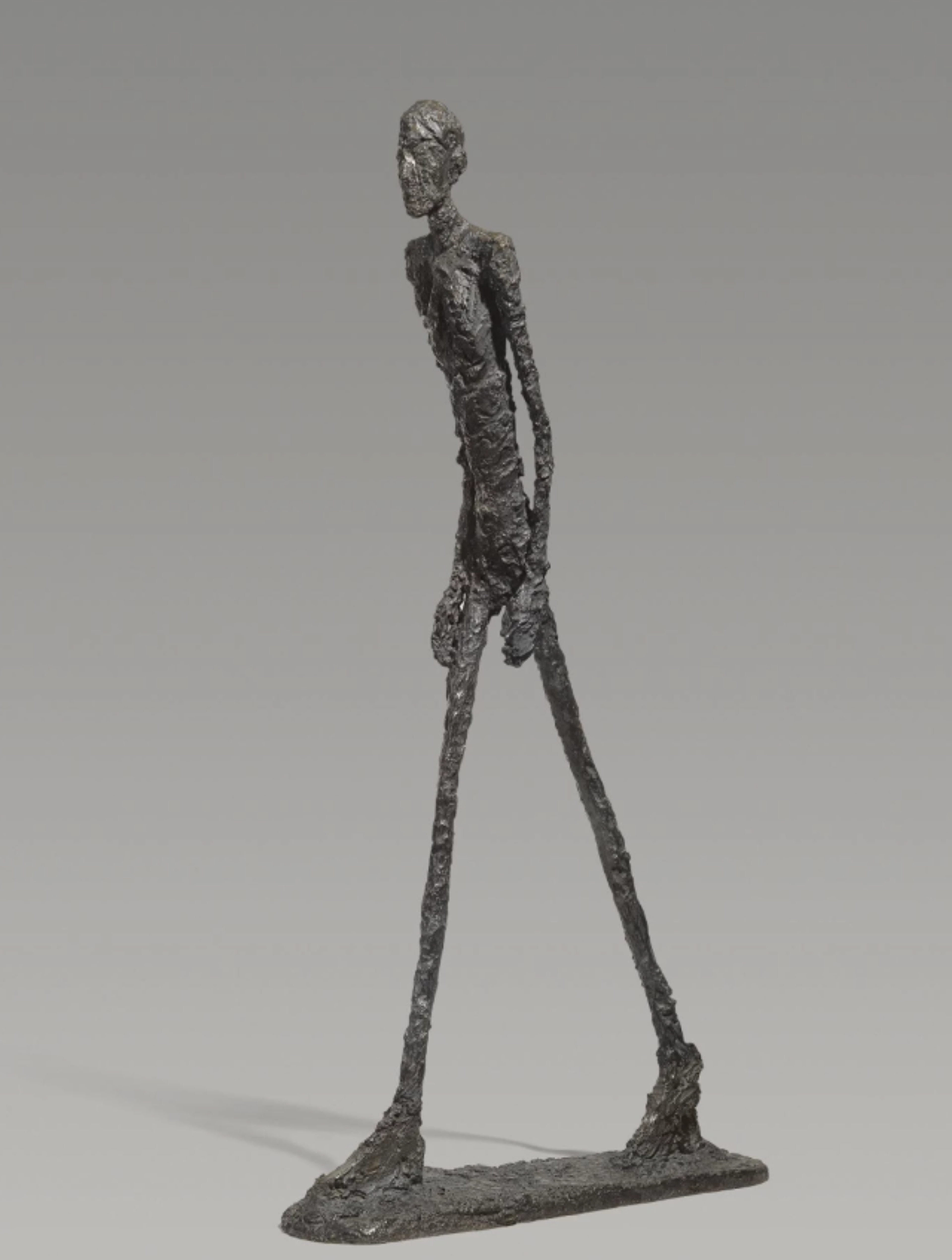 Tall bronze sculpture of an extremely thin, elongated figure leaning forwards in mid-stride. The figure has a rough, textured surface with long, stick-like legs and arms, creating a sense of forward movement despite its static medium.