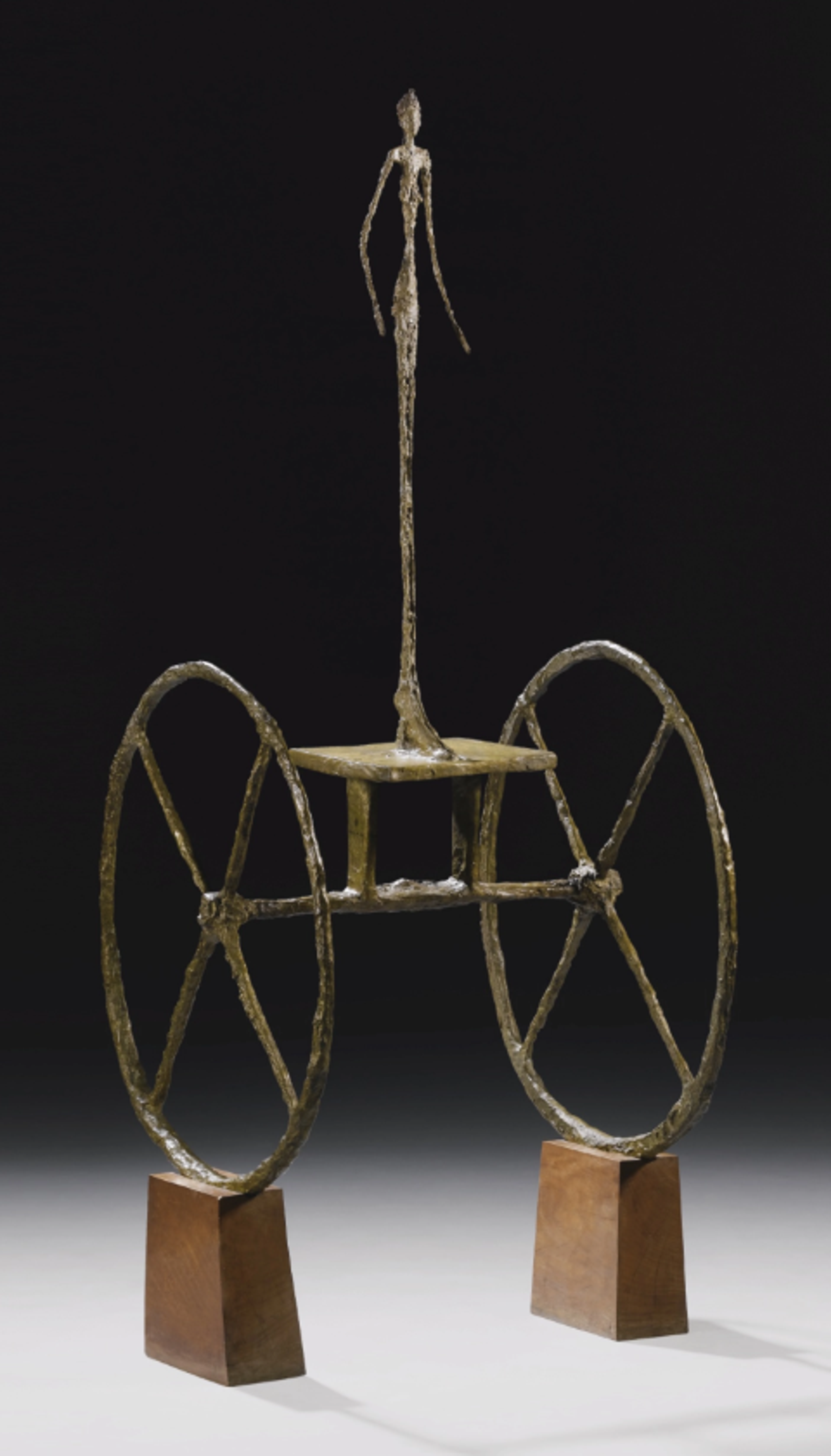 Bronze sculpture featuring a thin, vertical figure mounted on a two-wheeled cart. The large, spoked wheels contrast with the extremely slender central figure, all supported by two small wooden pedestals.
