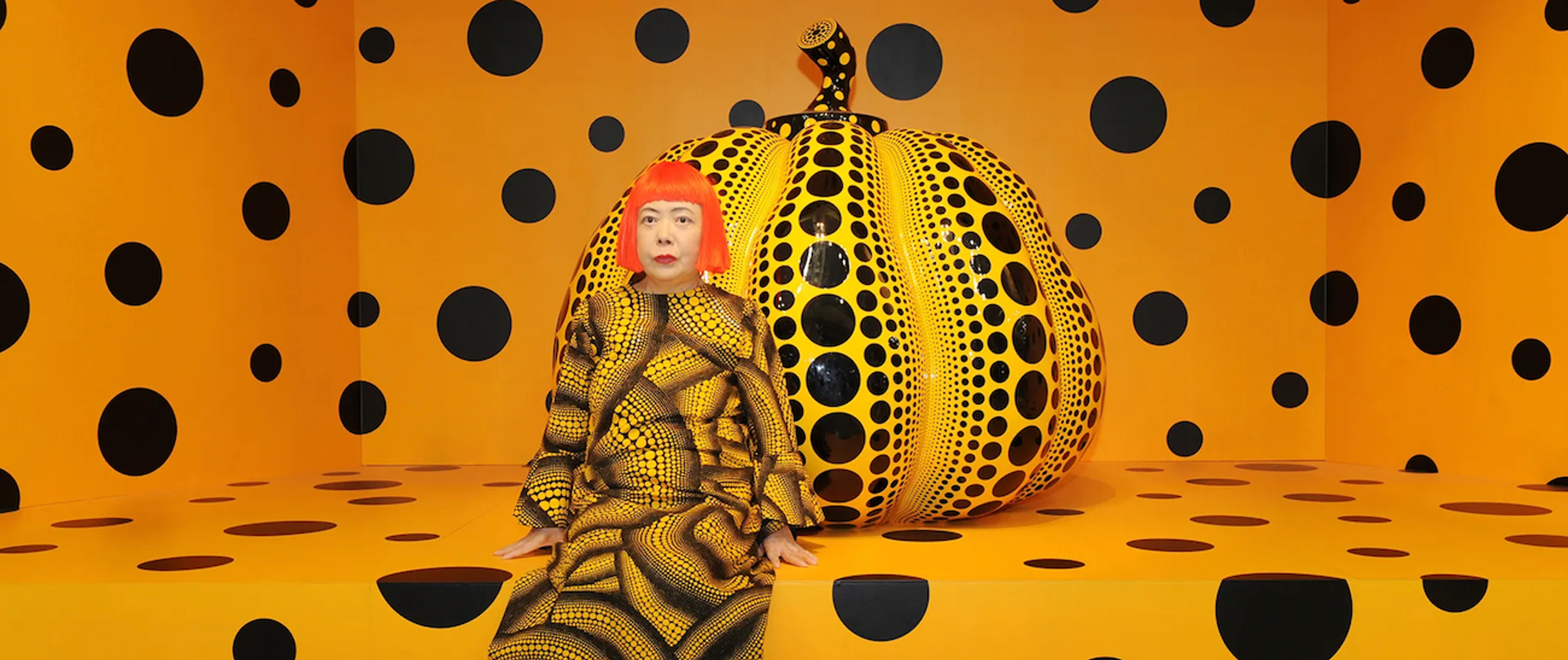 Yayoi Kusama était assise devant une citrouille grandeur nature jaune et noire dans une pièce orange recouverte de pois noirs.