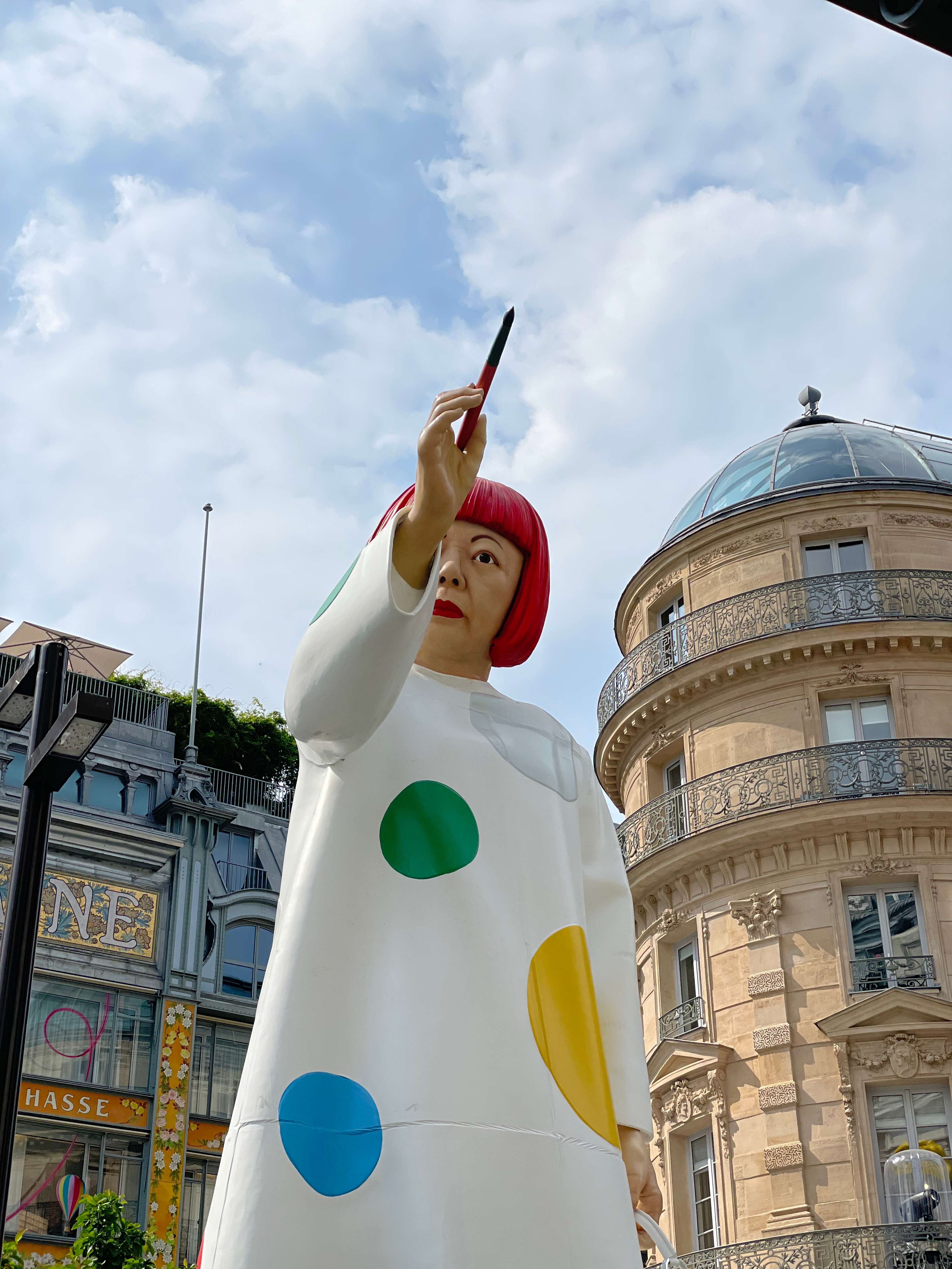 Une photographie de la grande sculpture de l'artiste Yayoi Kusama à Paris. La sculpture représente l'artiste coiffée d'une perruque rouge à la coupe au bol, vêtue d'une robe blanche à pois colorés, tenant un pinceau.
