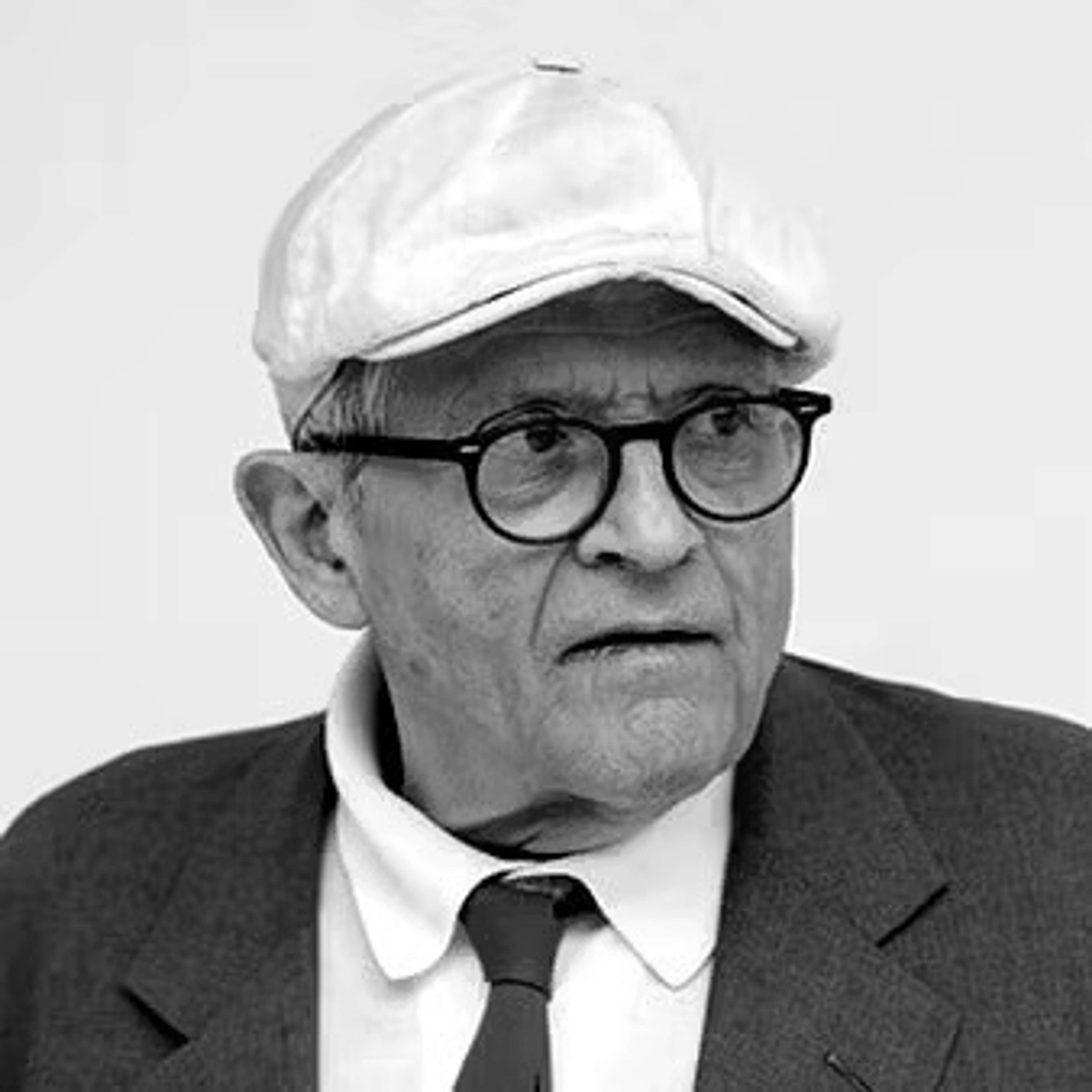 Black and white image of David Hockney wearing a suit, his glasses and white cap.