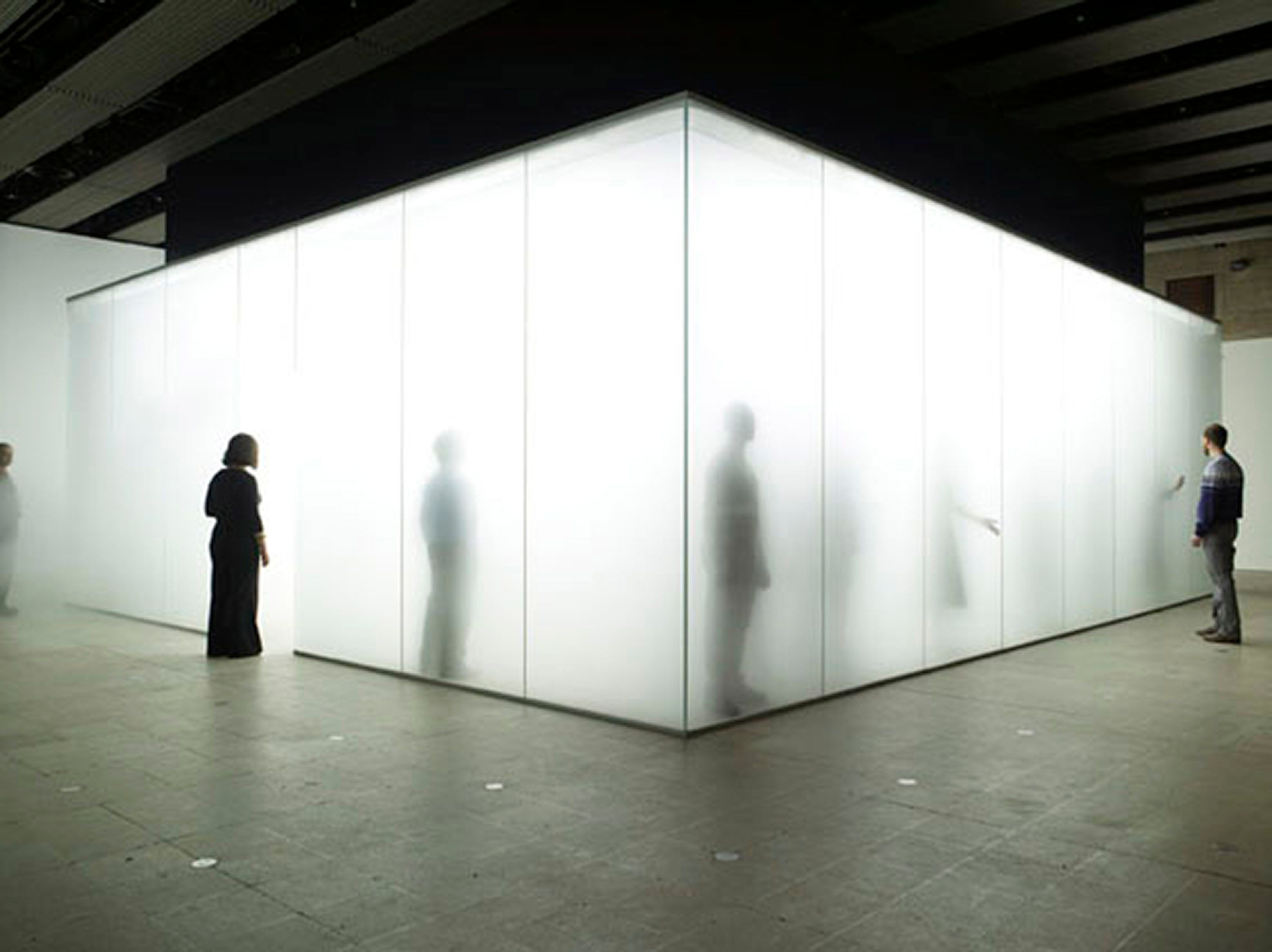 Cette photographie montre des personnes à l'intérieur d'une grande boîte lumineuse en verre remplie de brume. D'autres personnes se tiennent à l'extérieur de la boîte et observent.