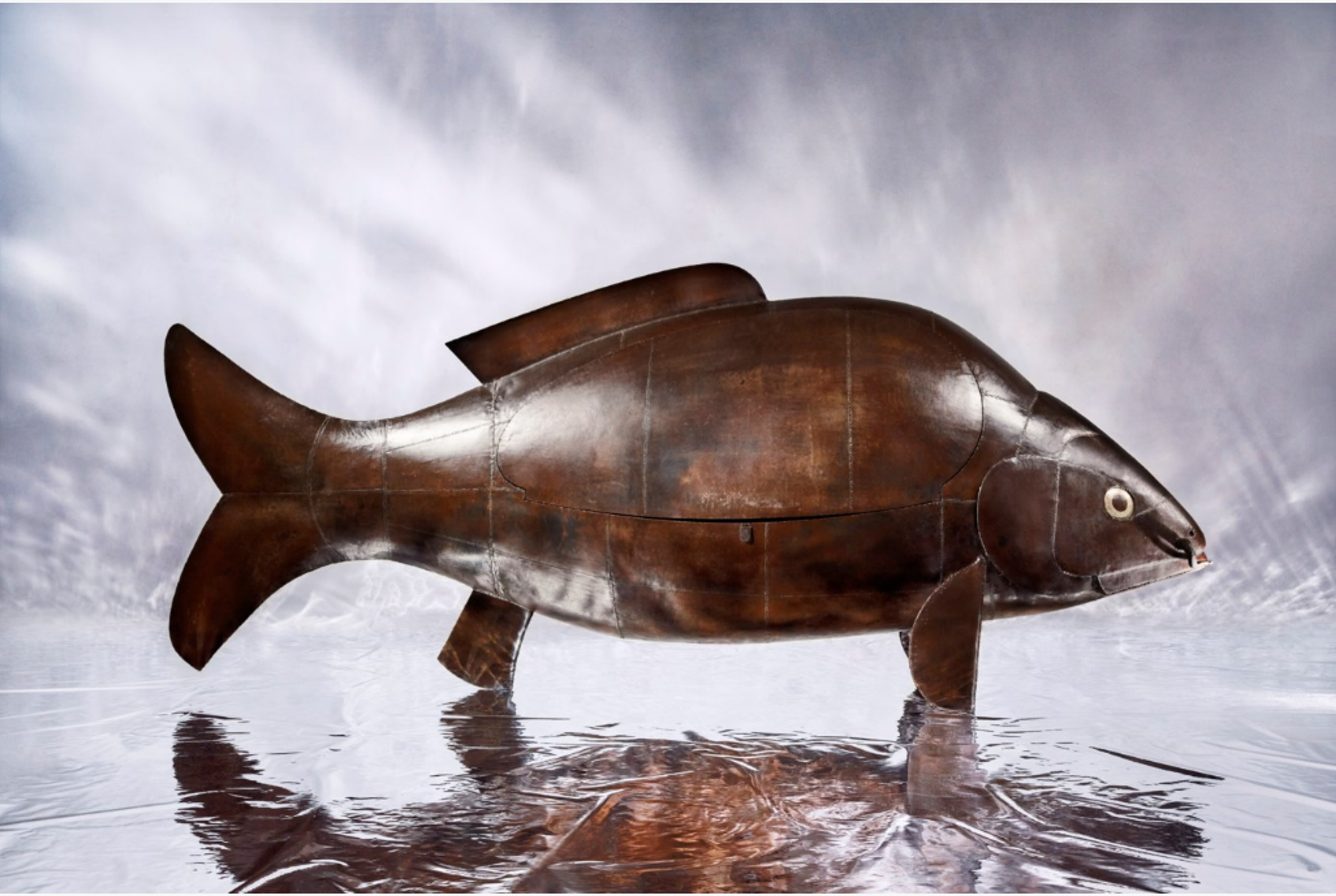 A metallic sculpture of a large fish (carp) with rich brown colouring. The sleek, curved form is assembled from sheets of iron, the joins of which are visible. Its overall shape is smooth with precise geometric edges. An oval shape on the carp’s side reveals where the bar opens. 