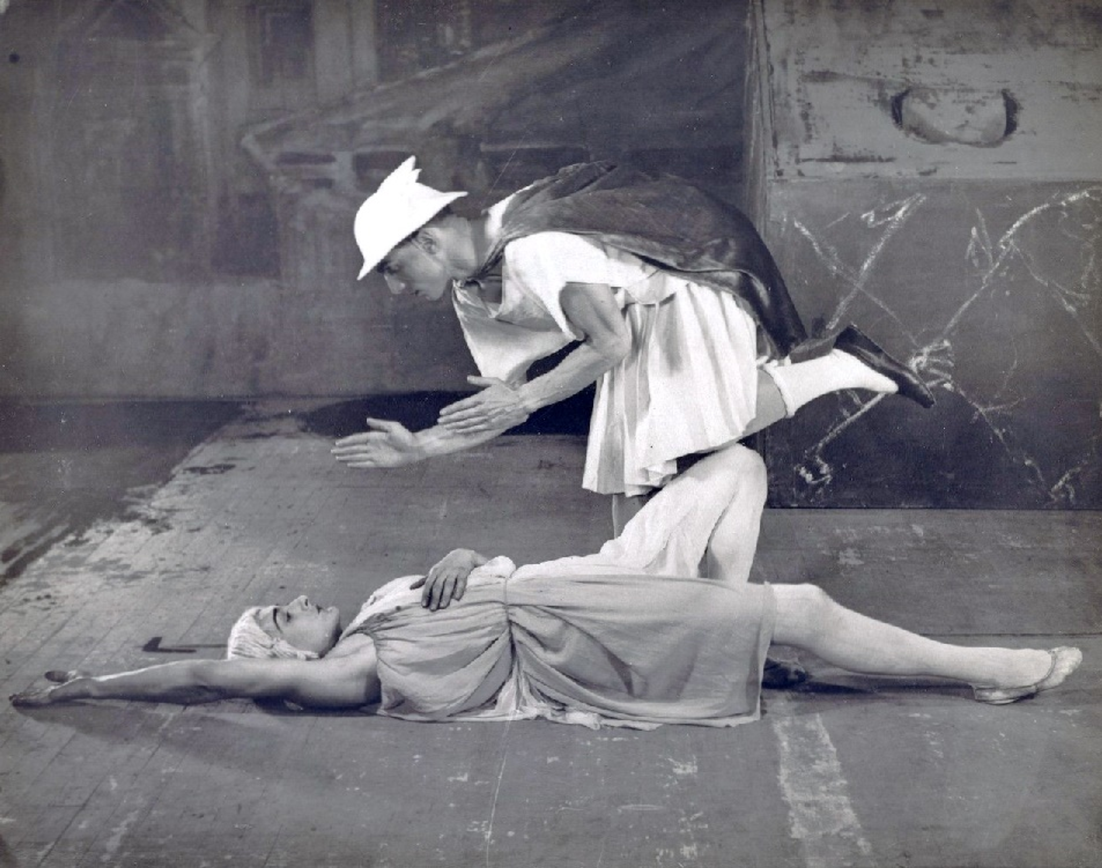 Une image monochrome montrant Massine en train d'être tuée par Mercure, ce dernier portant une tunique et son chapeau emblématique. Elle est représentée allongée sur le sol, les bras tendus au-dessus de sa tête.