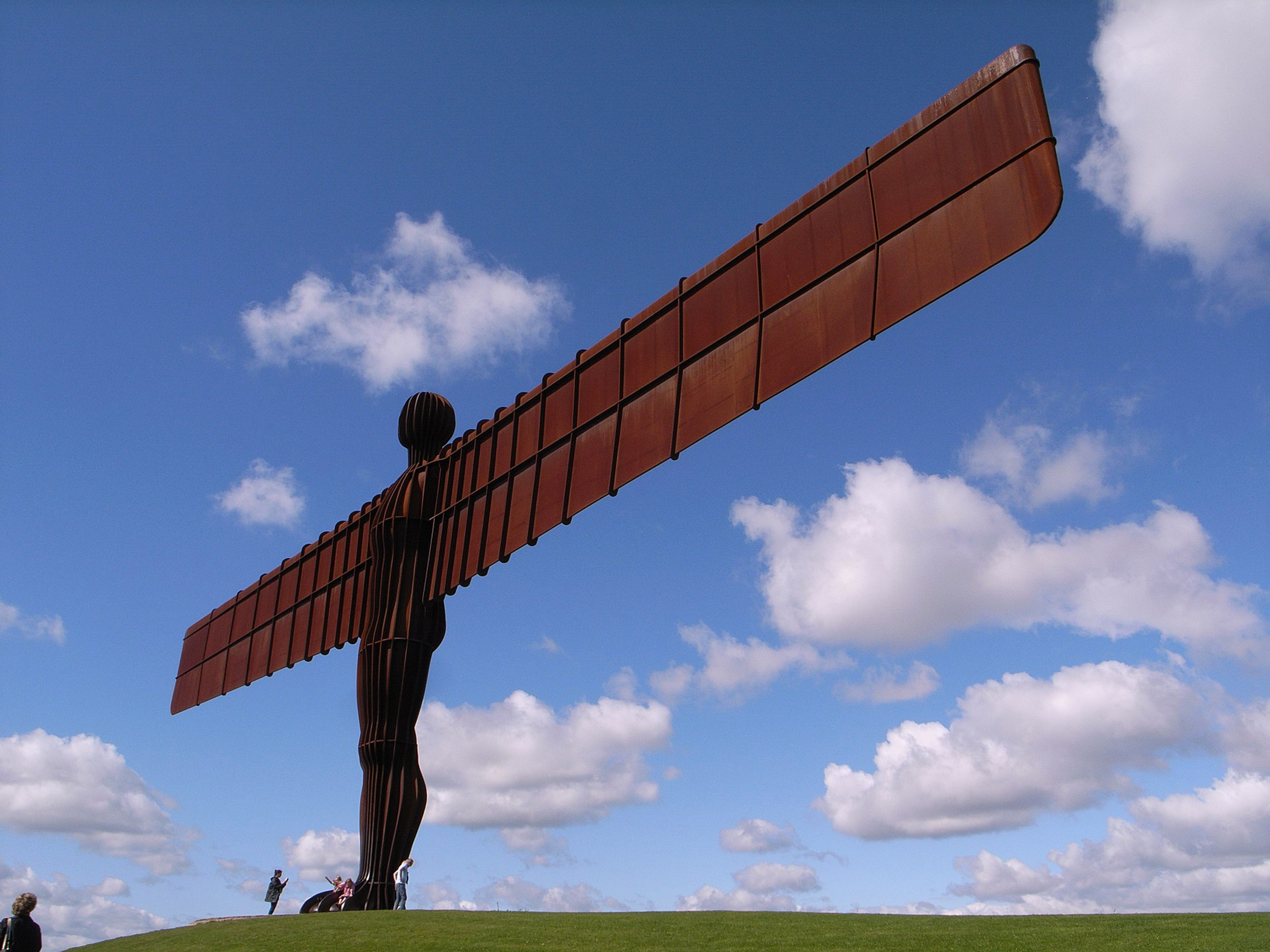 Photographie de la sculpture monumentale d'Antony Gormley, « Angel of the North », représentant une silhouette humaine aux ailes gigantesques lui servant de bras, se découpant sur un ciel ensoleillé.