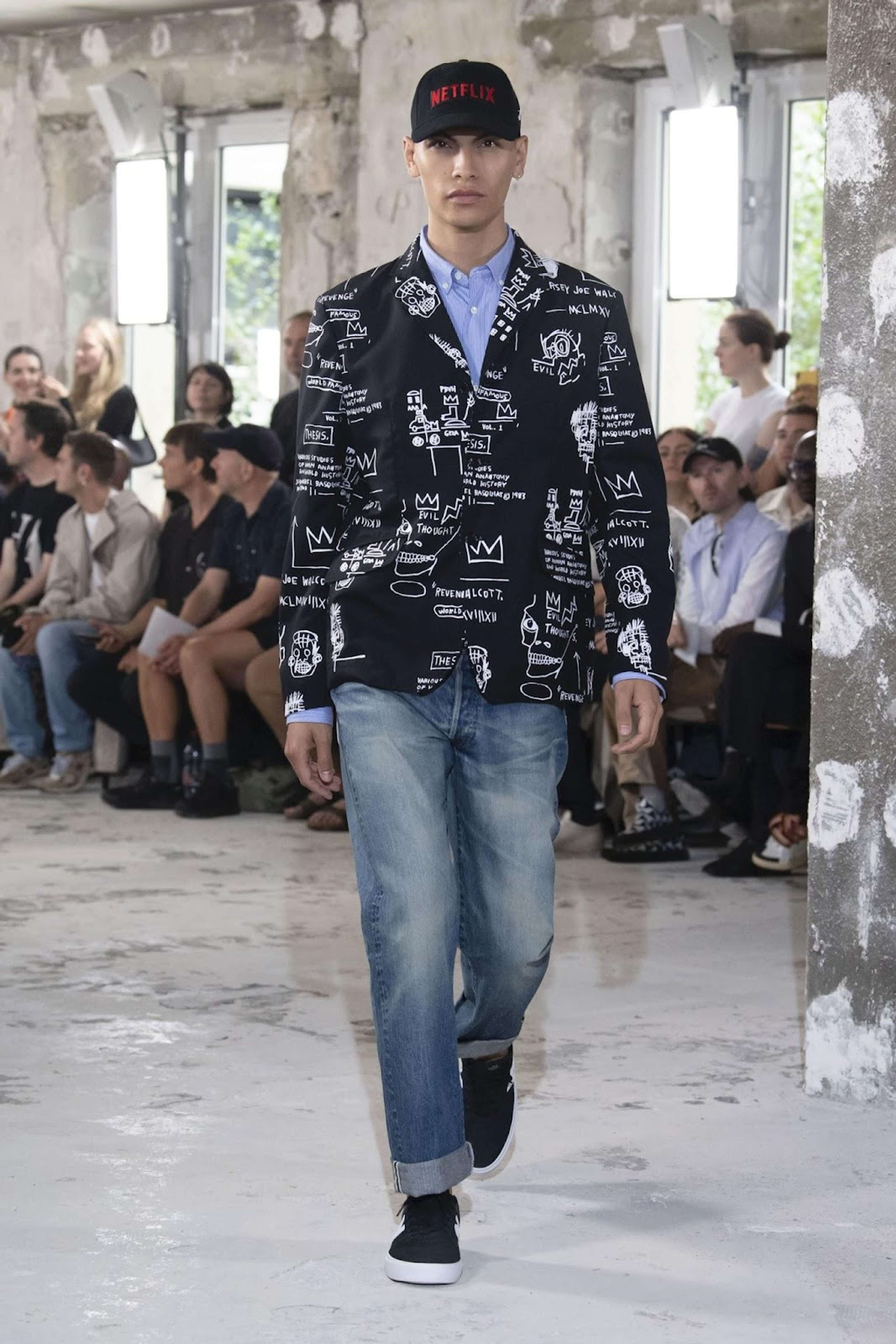 Une image d'un mannequin portant un costume noir Junya Watanabe griffé de motifs de Basquiat en blanc.