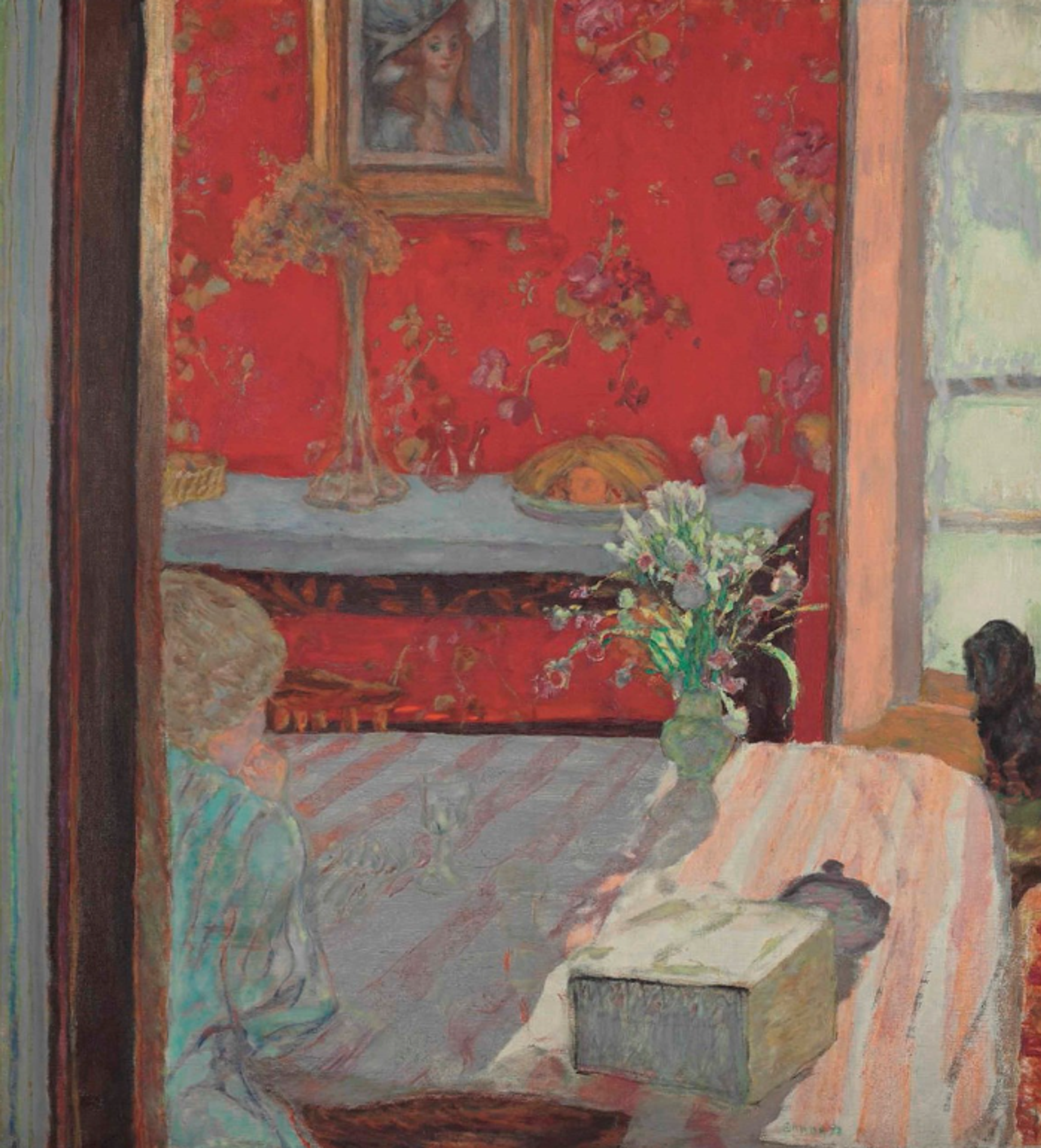 A red-walled interior with a seated figure just visible around a doorway, sat at a table with a pink-and-white striped tablecloth. The composition is rich with pattern and detail, showing part of Bonnard's Paris apartment.