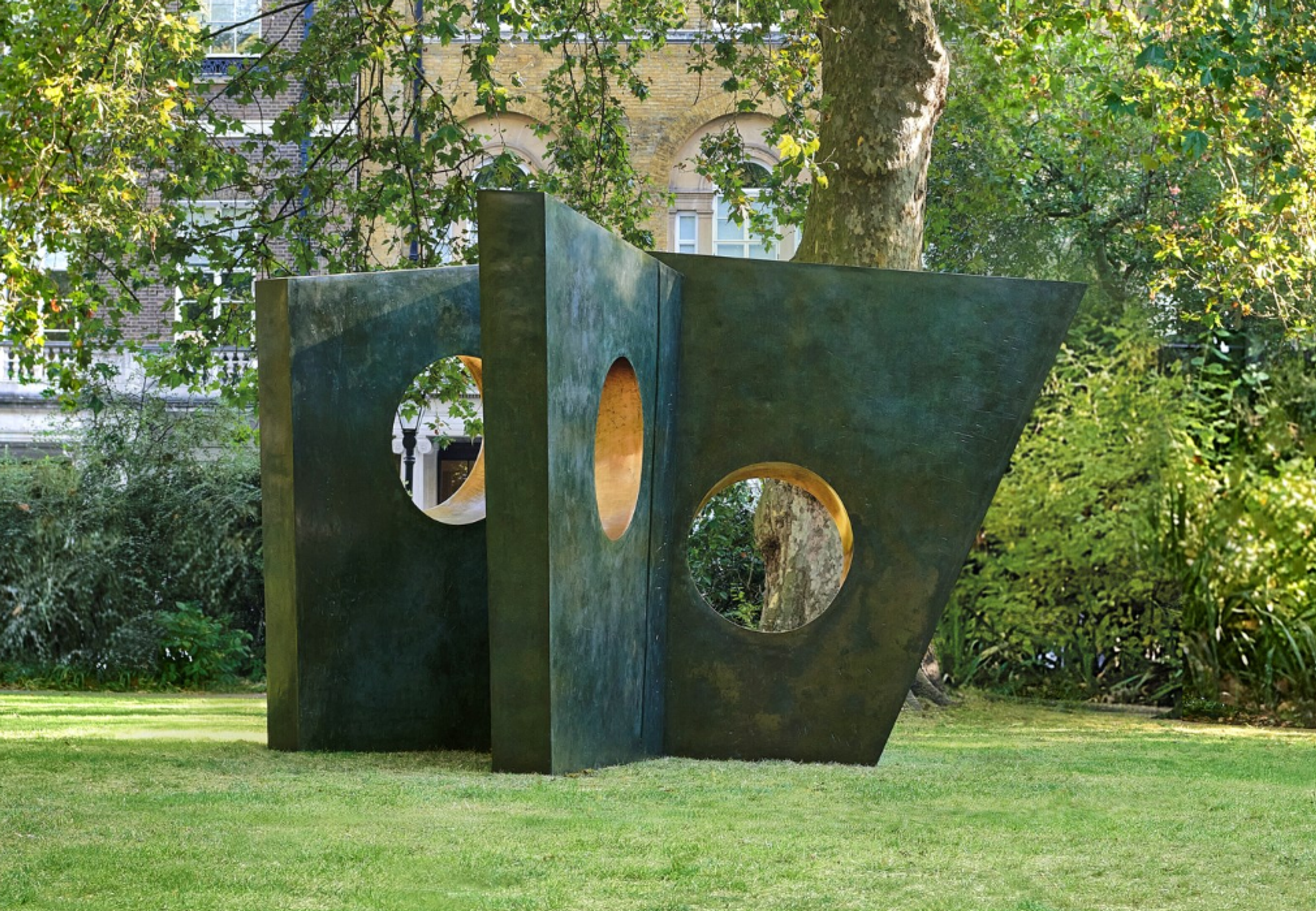 A large-scale outdoor installation consisting of angular bronze forms arranged to create a walkable environment. The structure features three geometric shapes with circular openings cut through them. Set in a garden with trees, the sculpture's dark bronze exterior contrasts with glimpses of sky and trees visible through its openings.