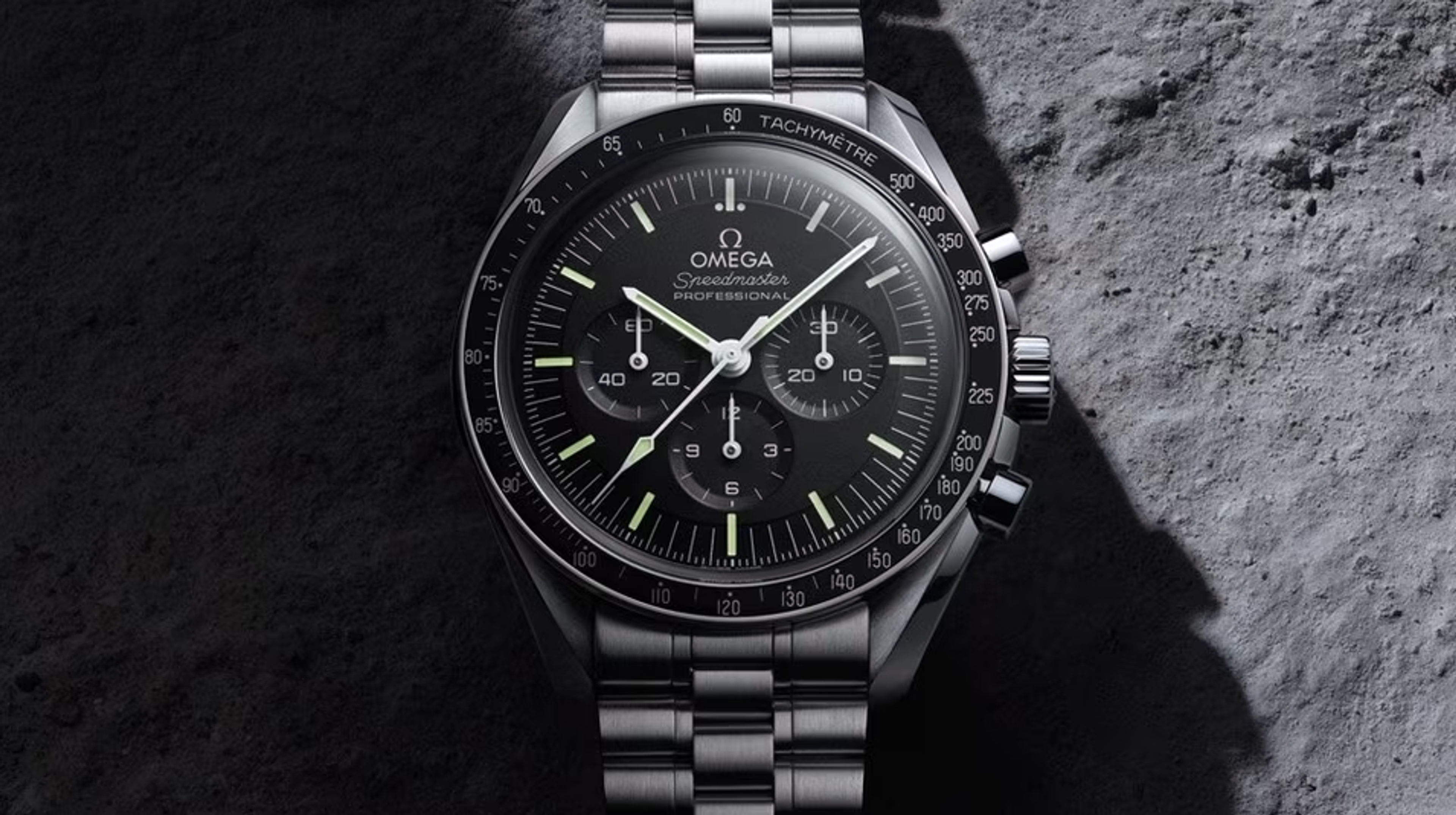 Une Omega Speedmaster noire, datant de 1957. Sur fond rocheux, le bracelet et le boîtier de la montre sont en métal argenté, avec un cadran noir et trois sous-cadrans.