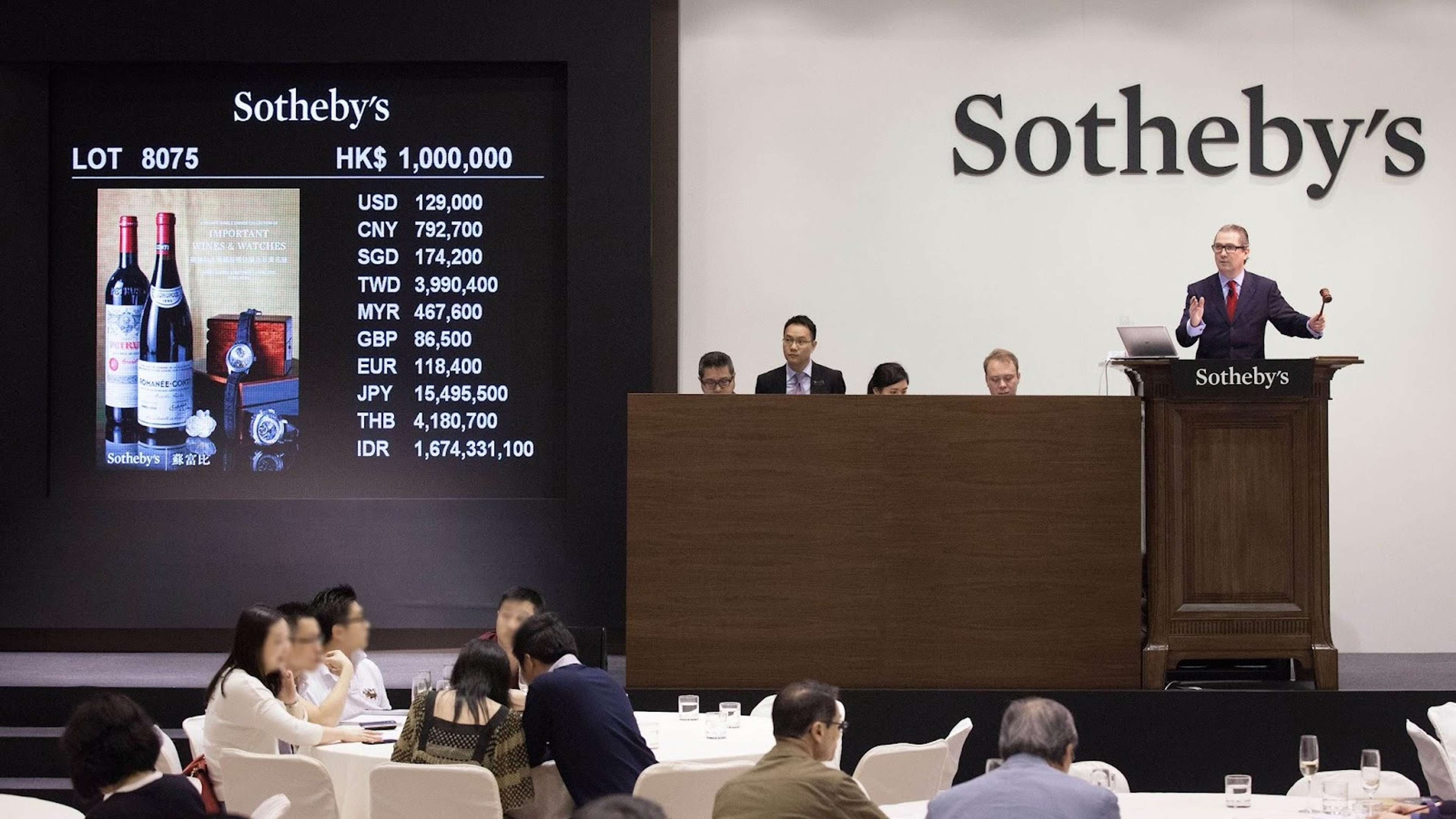 Salle de vente de Sotheby's, l'encanteur se tenant à droite de l'image avec son marteau, et l'image de gauche montrant un écran numérique affichant le lot de vin actuellement mis aux enchères.
